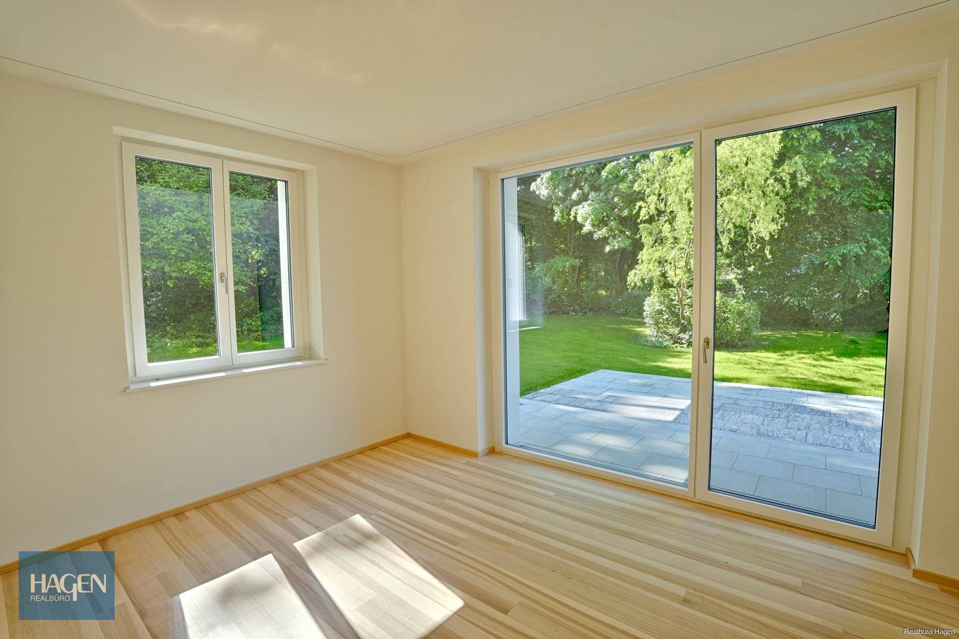 Leeres Zimmer mit Dielenboden, weißen Wänden und großen Glasschiebetüren zum Gartenbereich.
