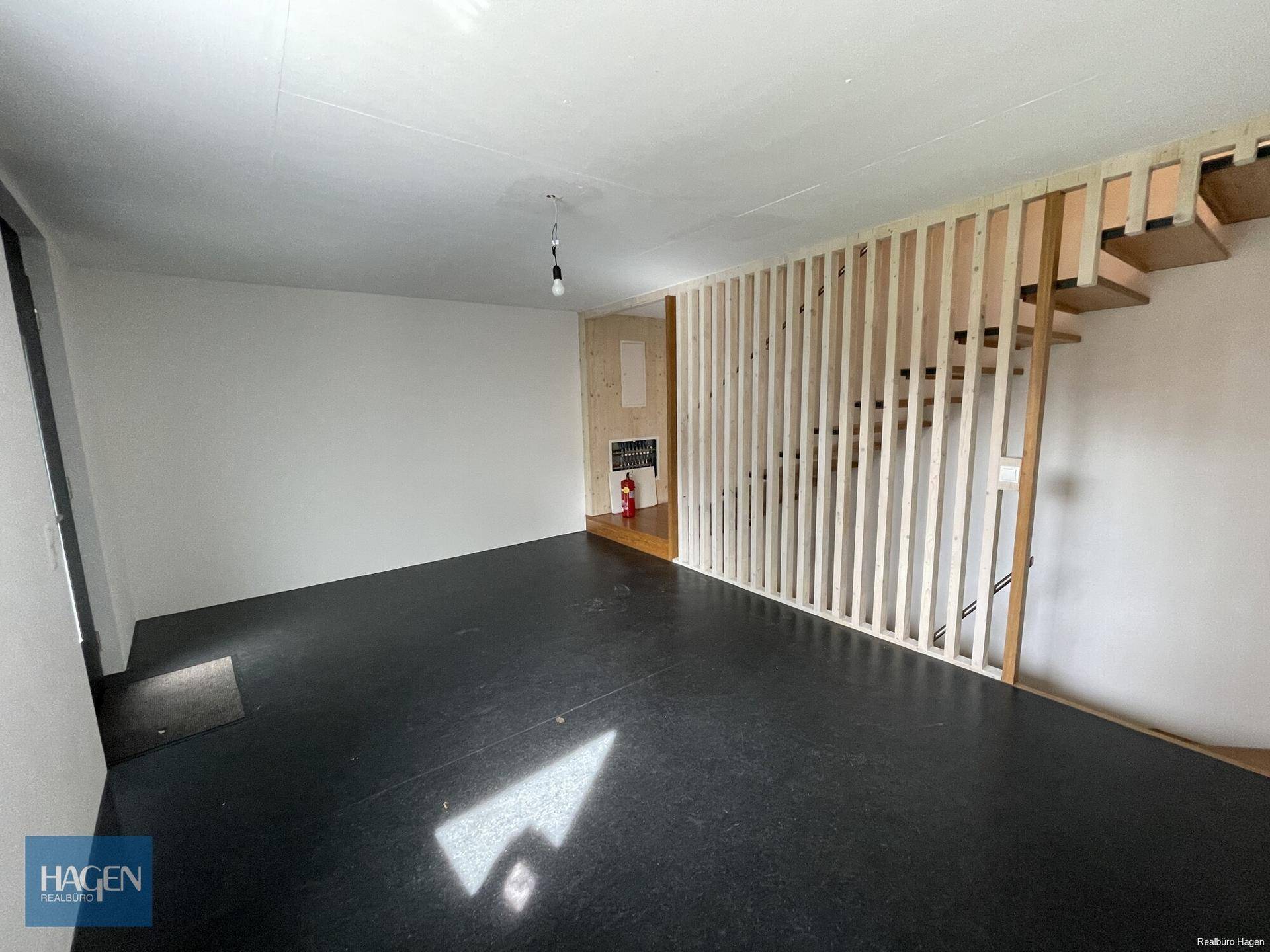 Heller, leerer Raum mit dunklem Boden und offener Holztreppe, die in die obere Etage führt.