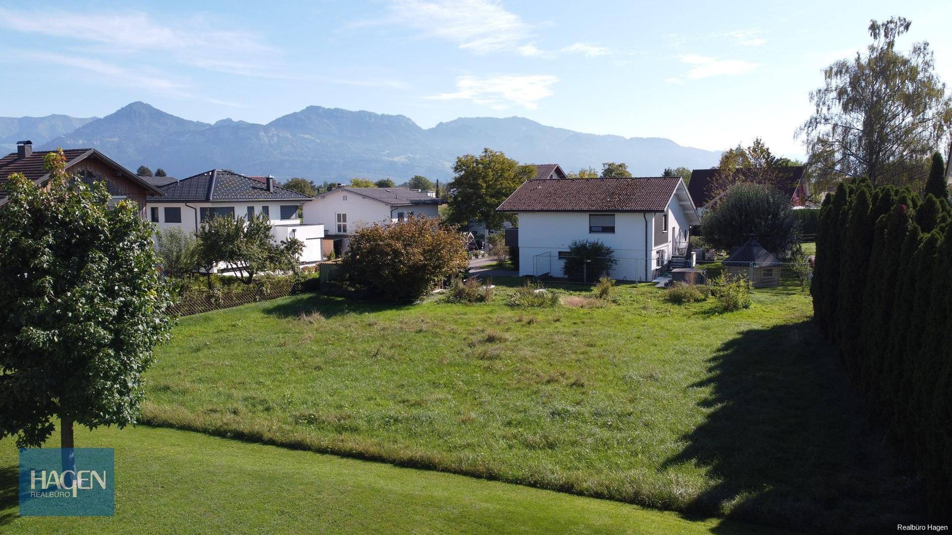 Weitläufiges Wiesengrundstück mit Bergblick und angrenzender Wohnbebauung unter blauem Himmel.
