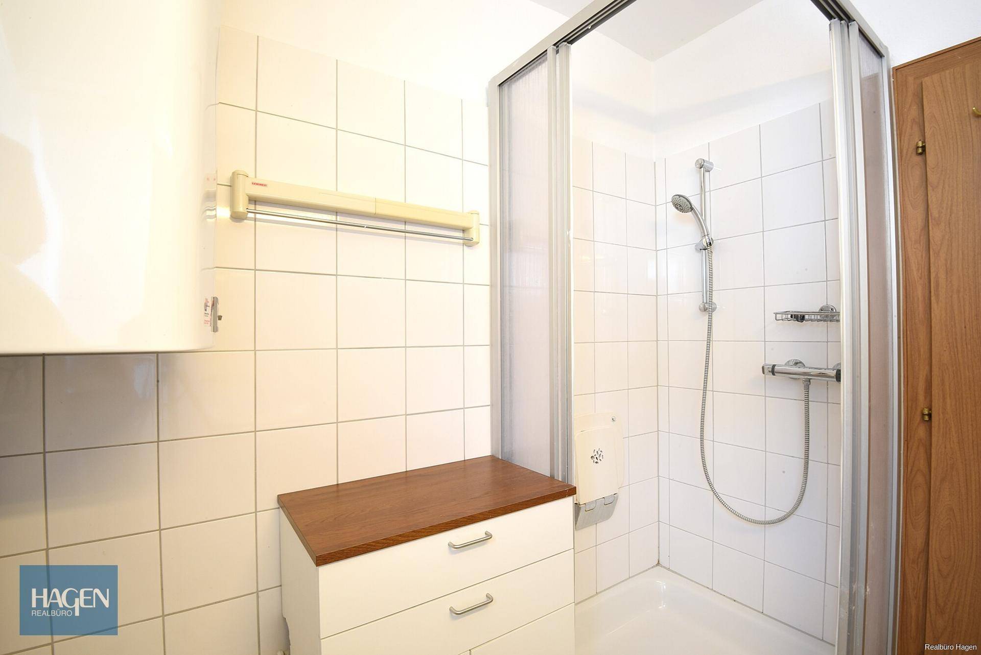 Badezimmer mit weiß gefliesten Wänden, einer Duschkabine und einem kleinen Waschtischunterschrank.