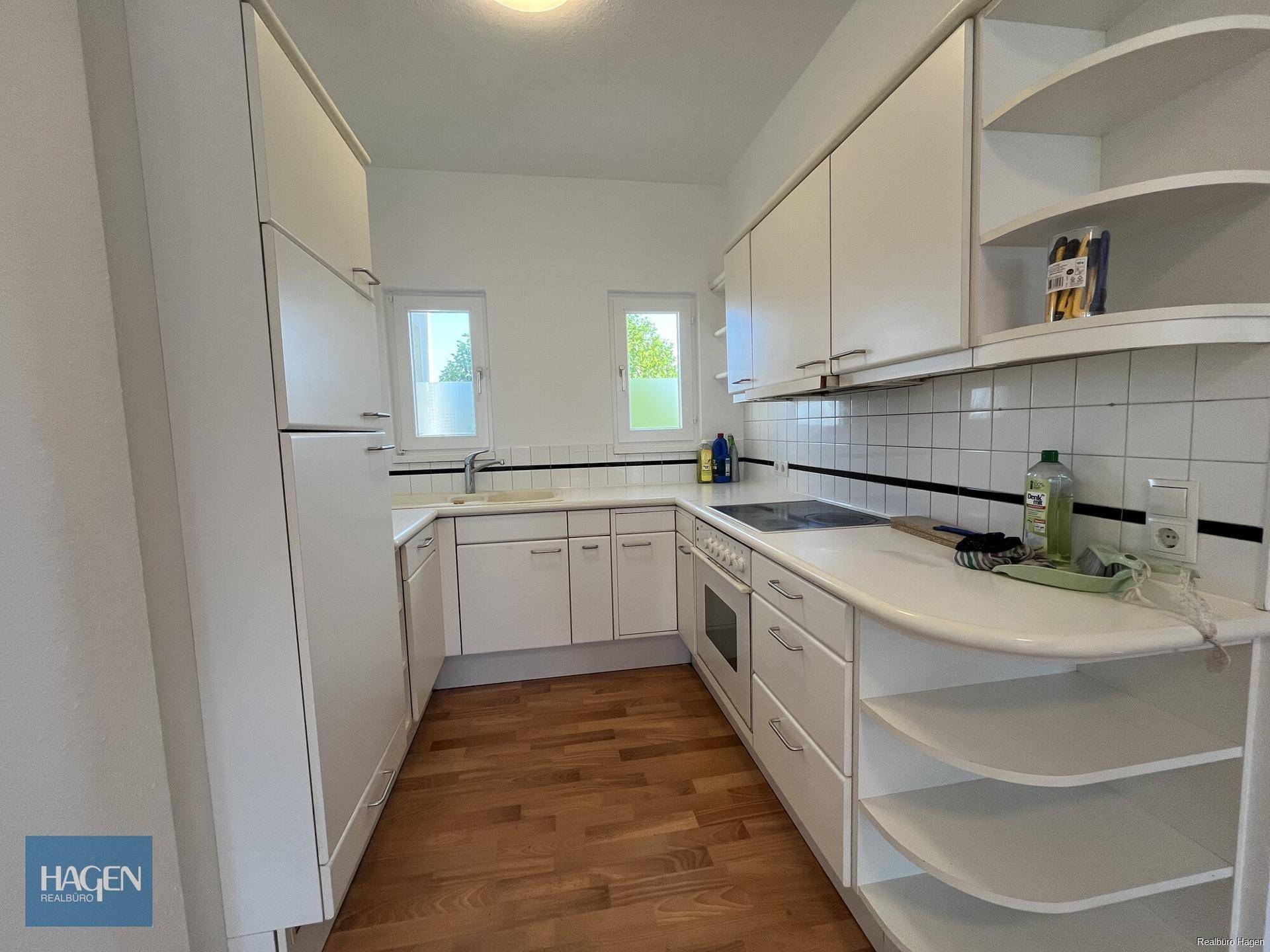Helle Küche mit weißen Schränken, Laminatboden und zwei Fenstern für Tageslicht.
