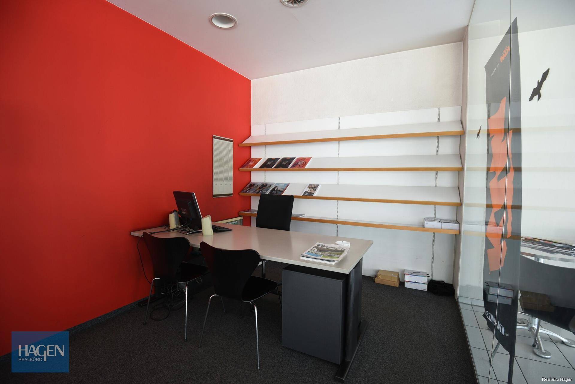 Kleiner Bürobereich mit einer roten Akzentwand, einem Schreibtisch und Regalen für Broschüren.