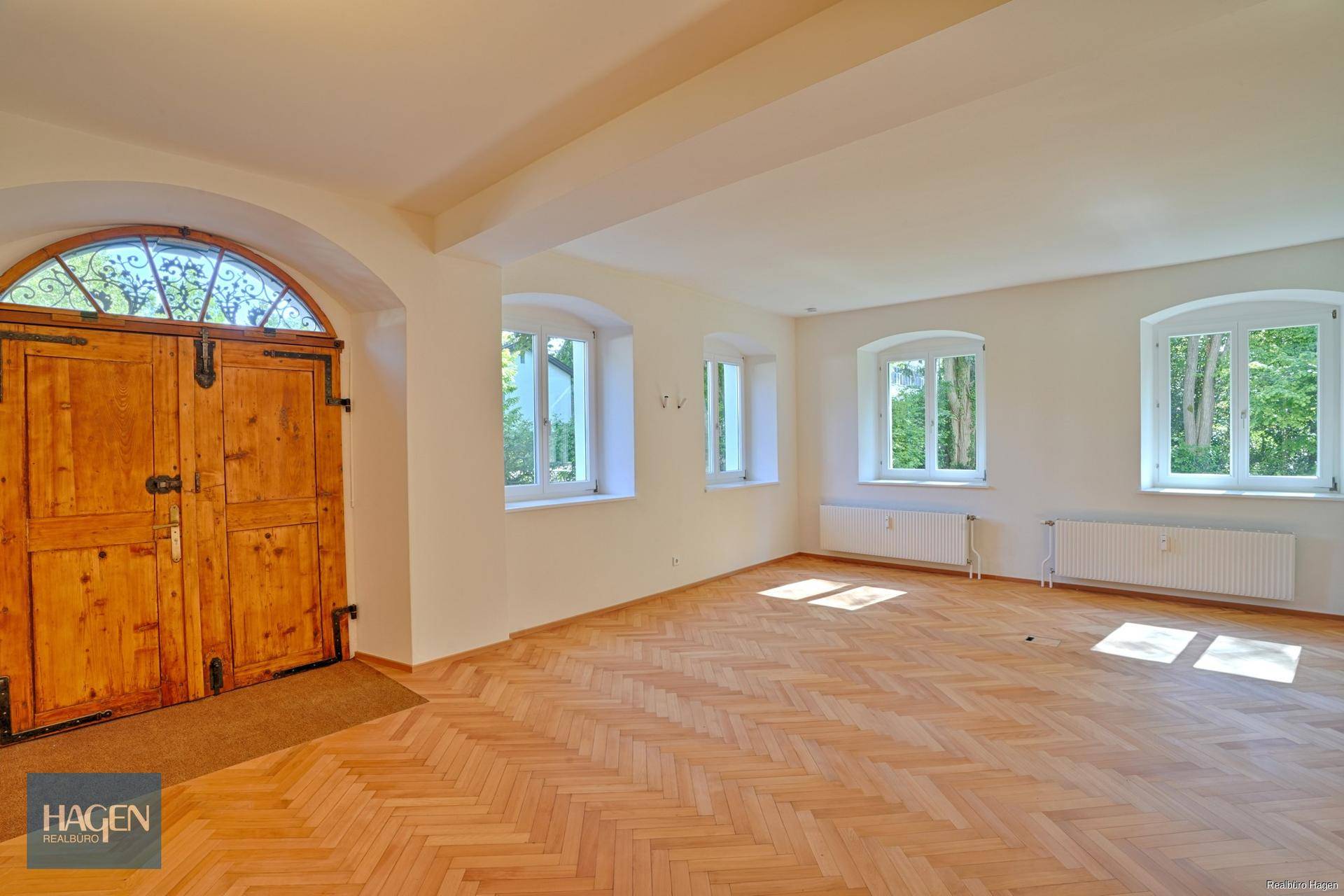 Geräumiger Innenraum mit Fischgrätparkett, weißen Wänden, mehreren Fenstern und einer großen Holztür.