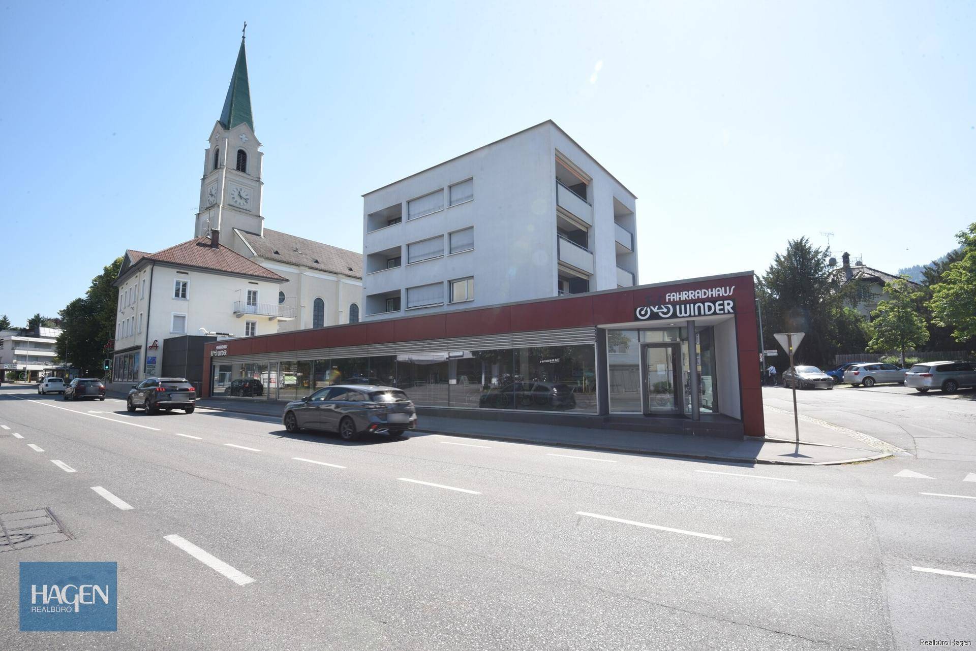 Weitwinkelaufnahme des Geschäftsgebäudes mit angrenzender Straße und einem Kirchturm im Hintergrund.