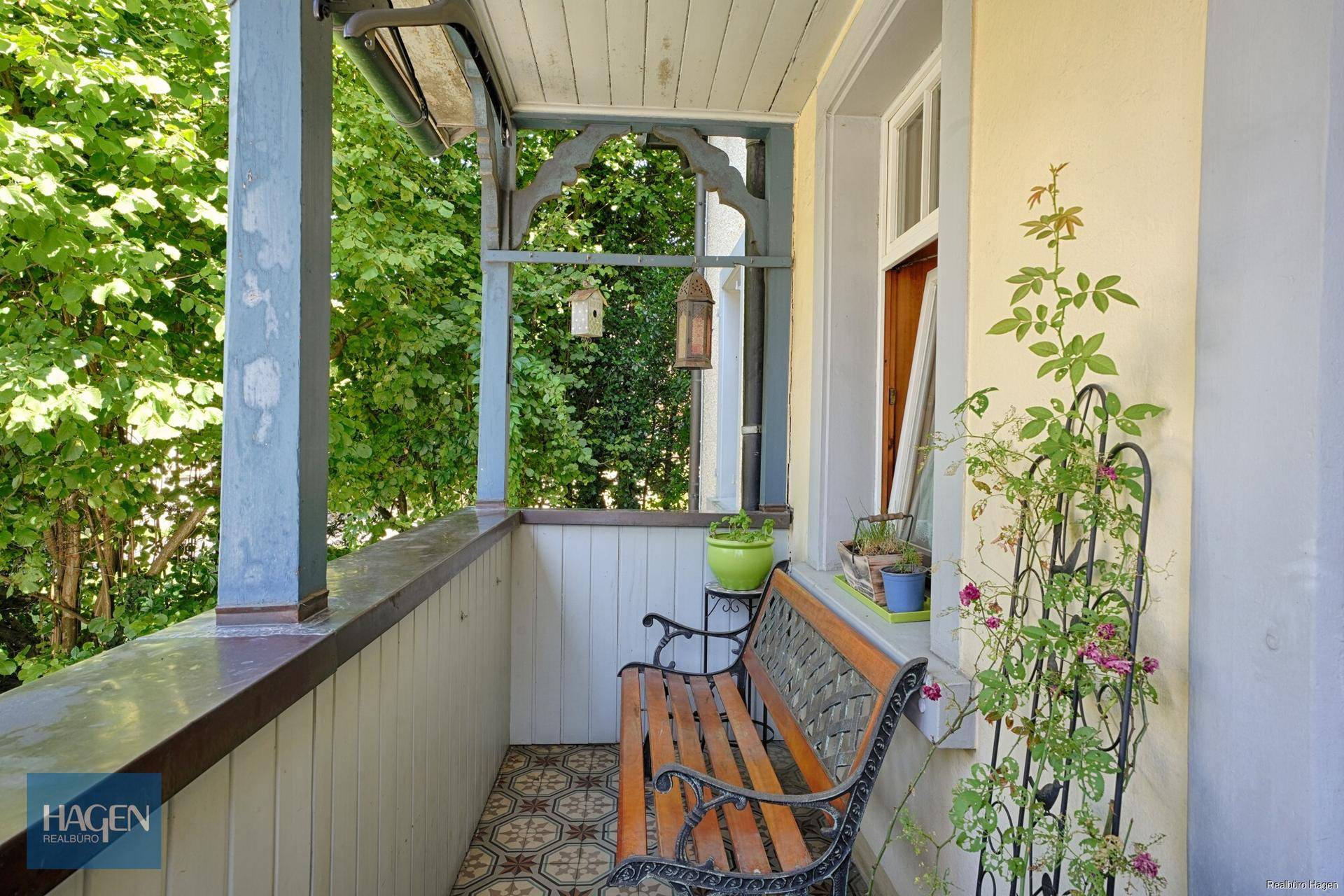 Kleiner überdachter Balkon mit gemusterten Fliesen, Holzbank und Blick ins Grüne.