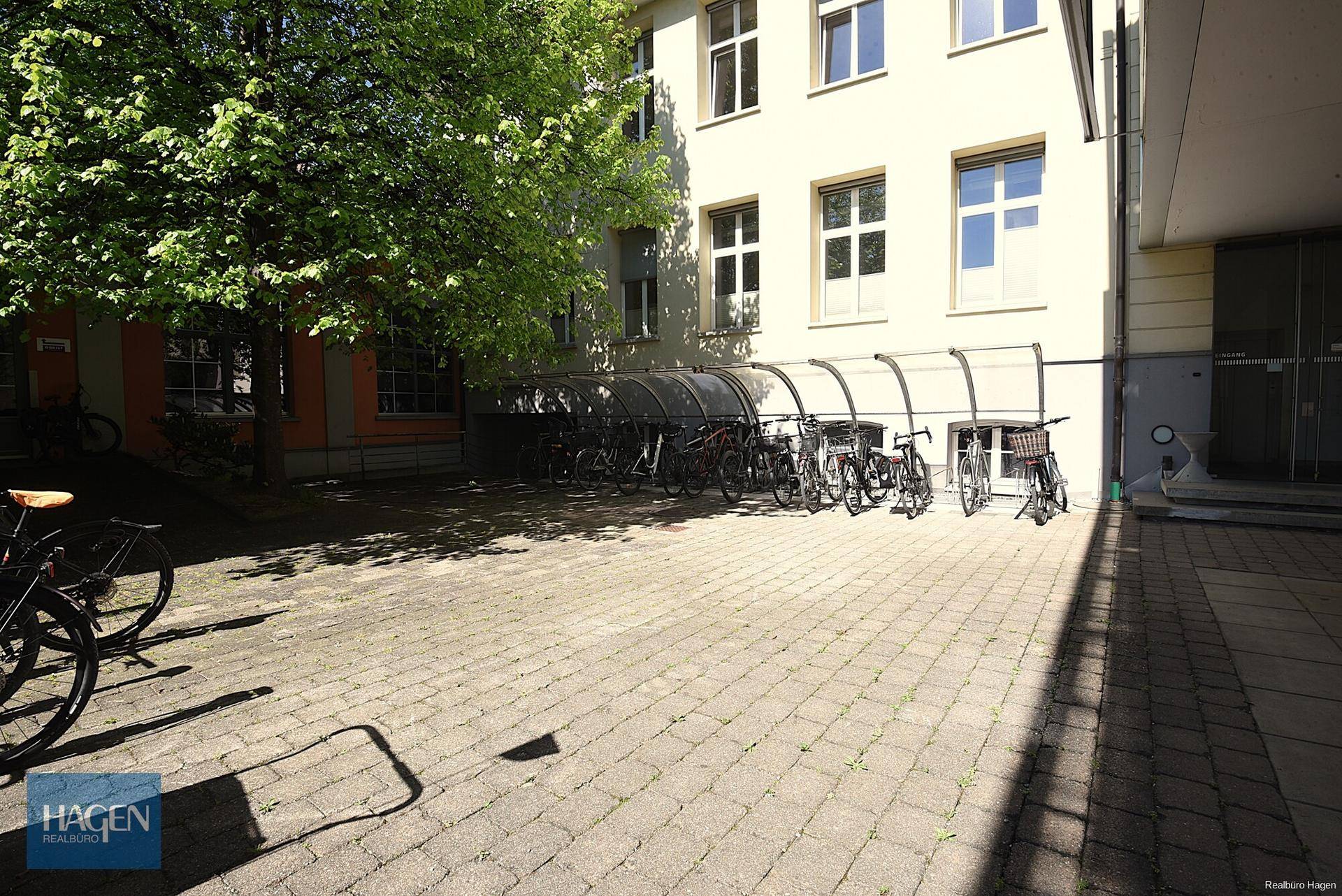Überdachter Fahrradabstellplatz im Freien neben dem Gebäude an einem sonnigen Tag mit viel Grün.