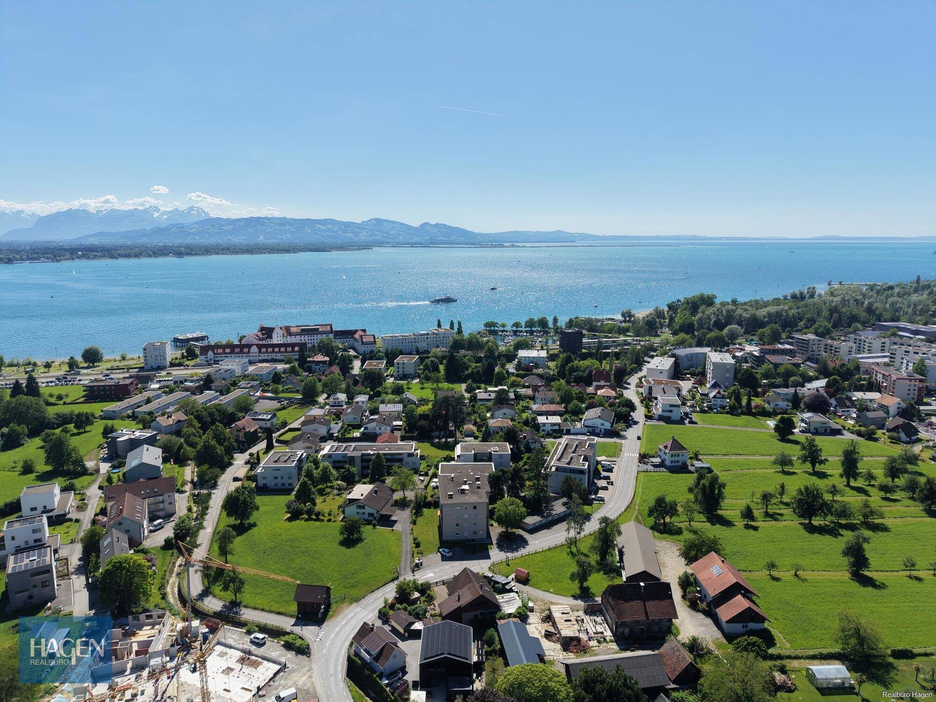 Luftaufnahme einer Küstenstadt am See mit Bergpanorama und Bauarbeiten im Vordergrund.