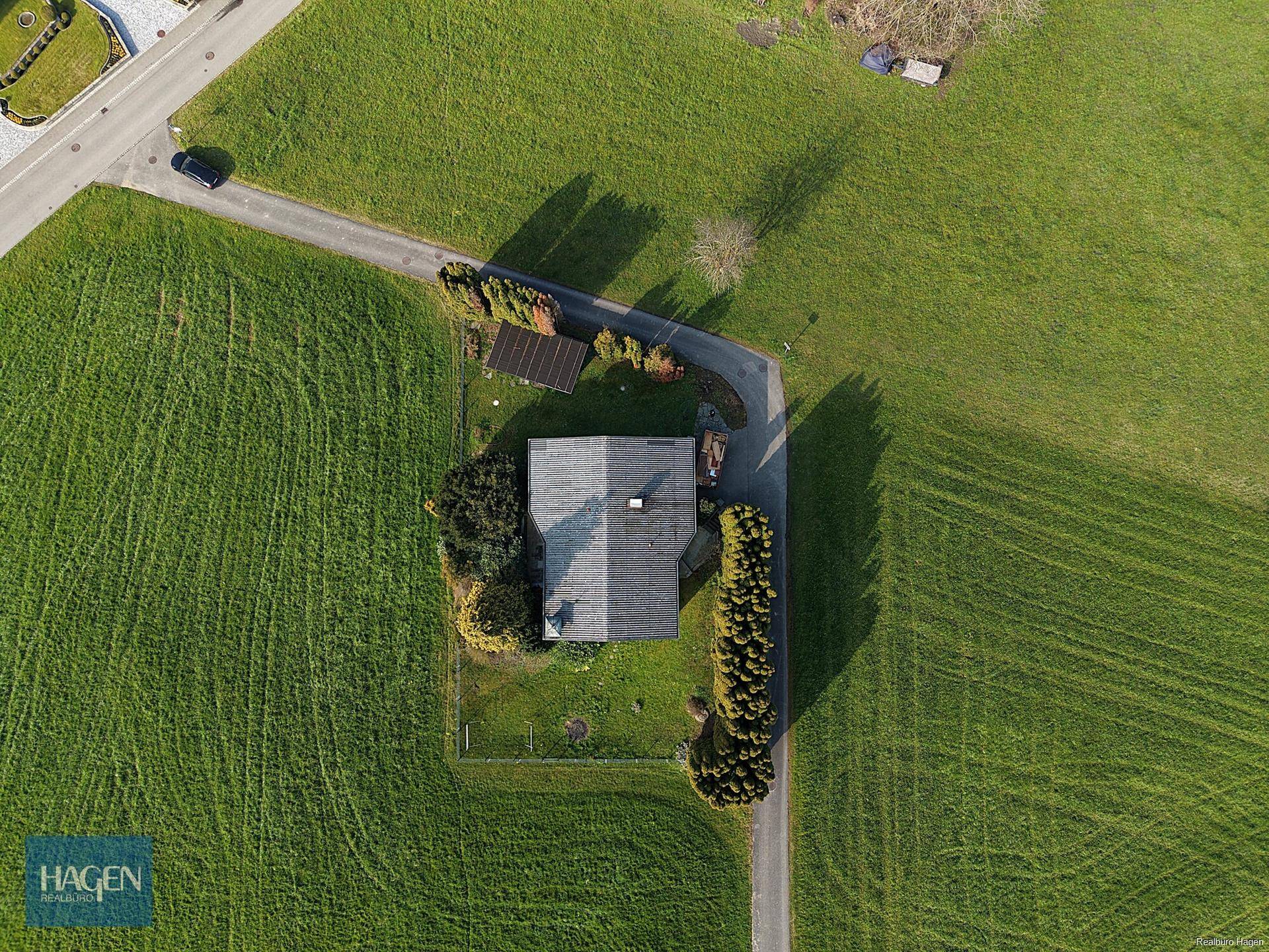 Luftaufnahme eines Einfamilienhauses umgeben von grünen Wiesen und einer Zufahrtsstraße.