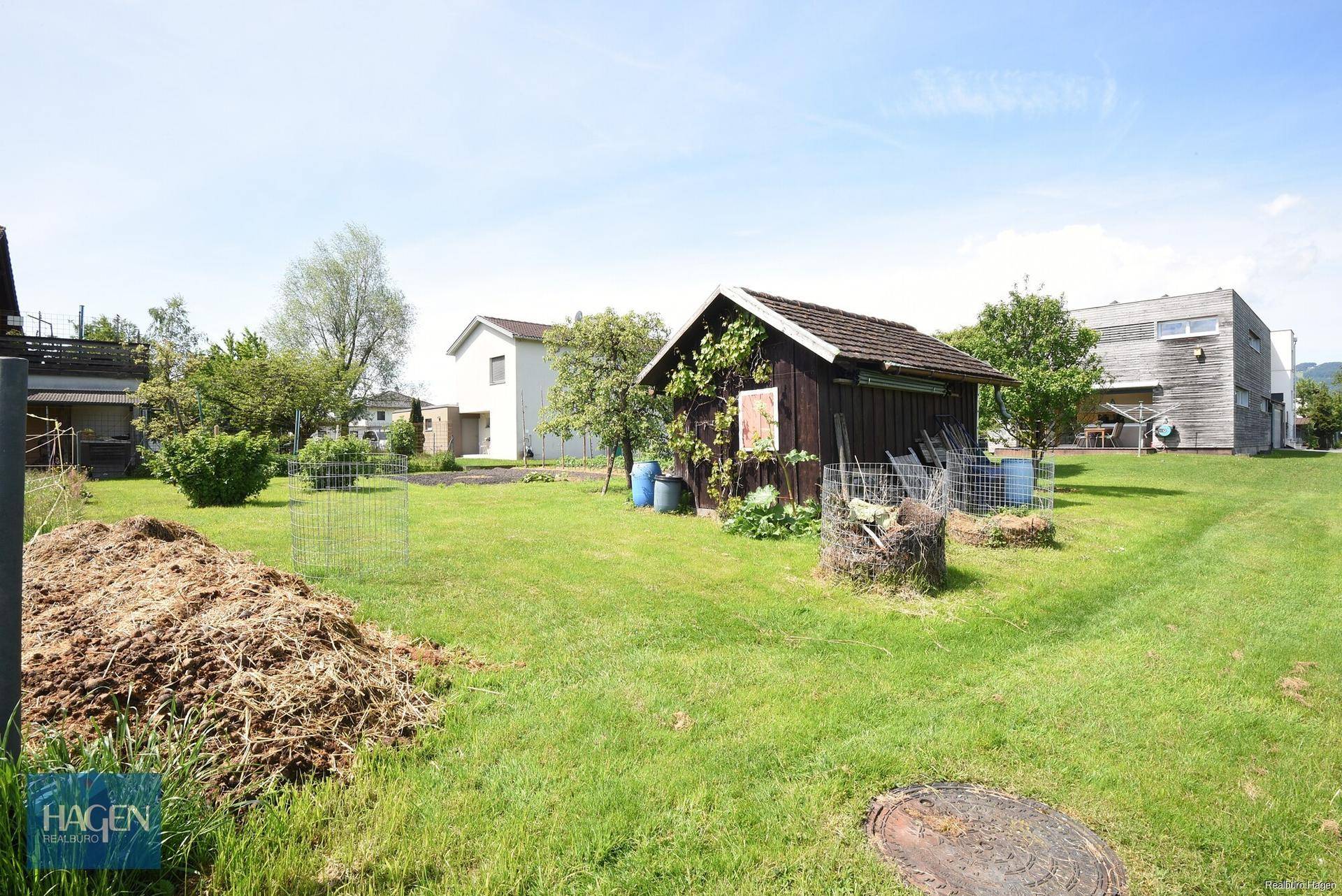 Gartenbereich mit einem kleinen Holzschuppen, umgeben von Rasenflächen und einigen Bäumen an einem sonnigen Tag.