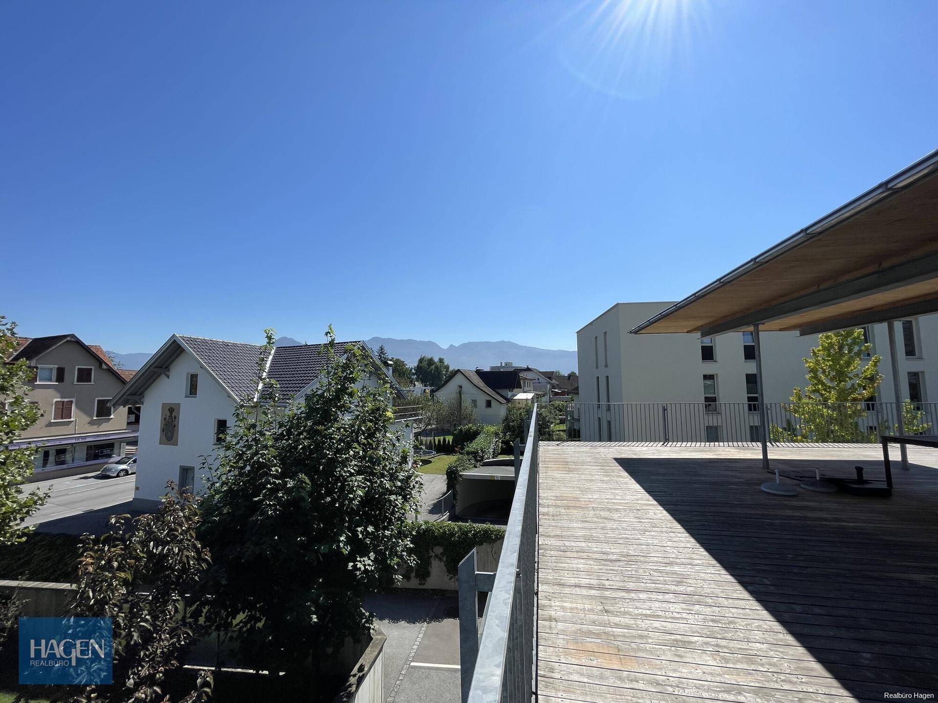 Sonniger Tag auf der Holzterrasse mit Blick auf die umliegende Wohnbebauung und Berge.