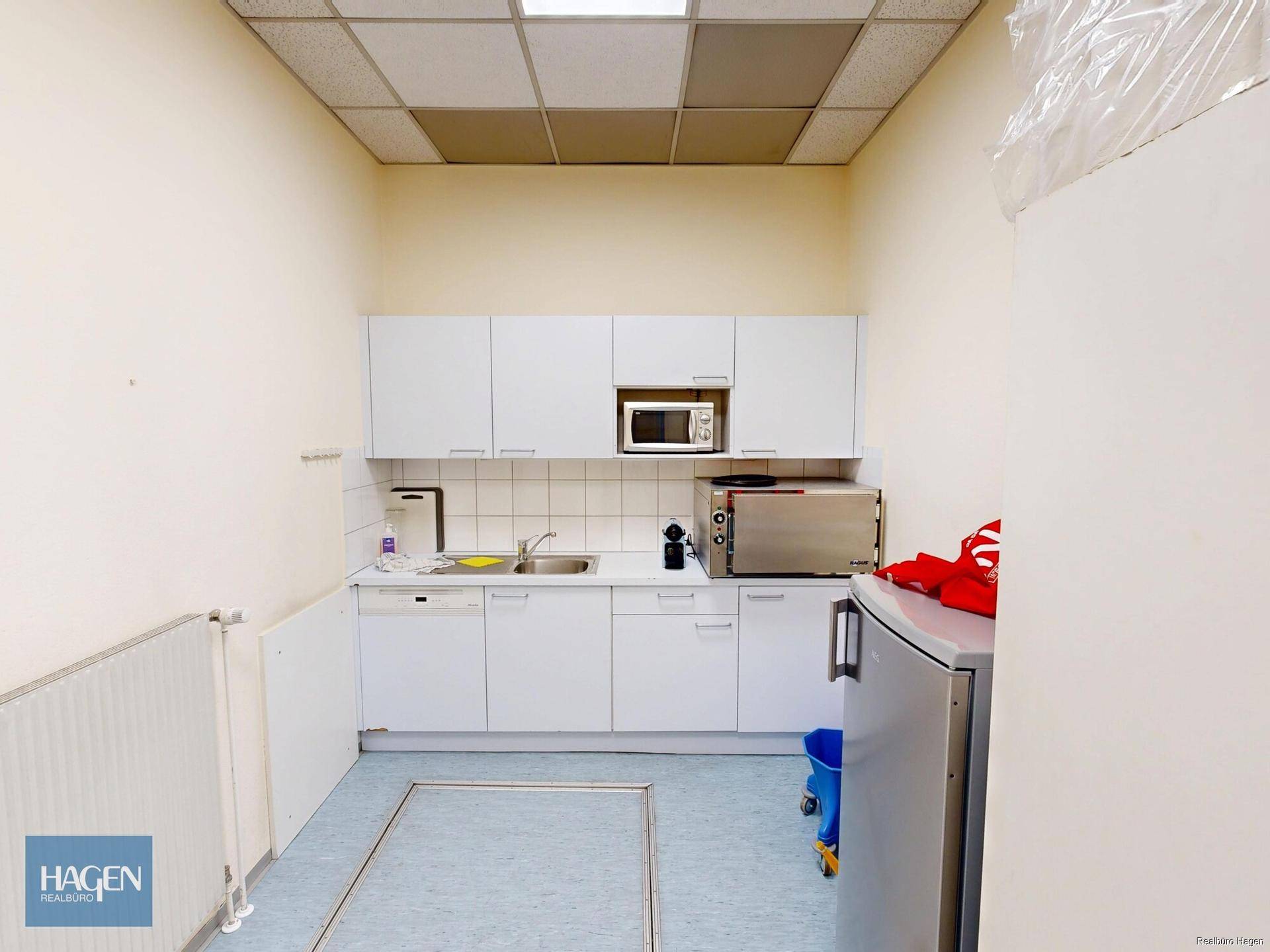 Kleine Küchenzeile mit weißen Schränken, Spüle, Mikrowelle und Kühlschrank in einem Pausenraum.