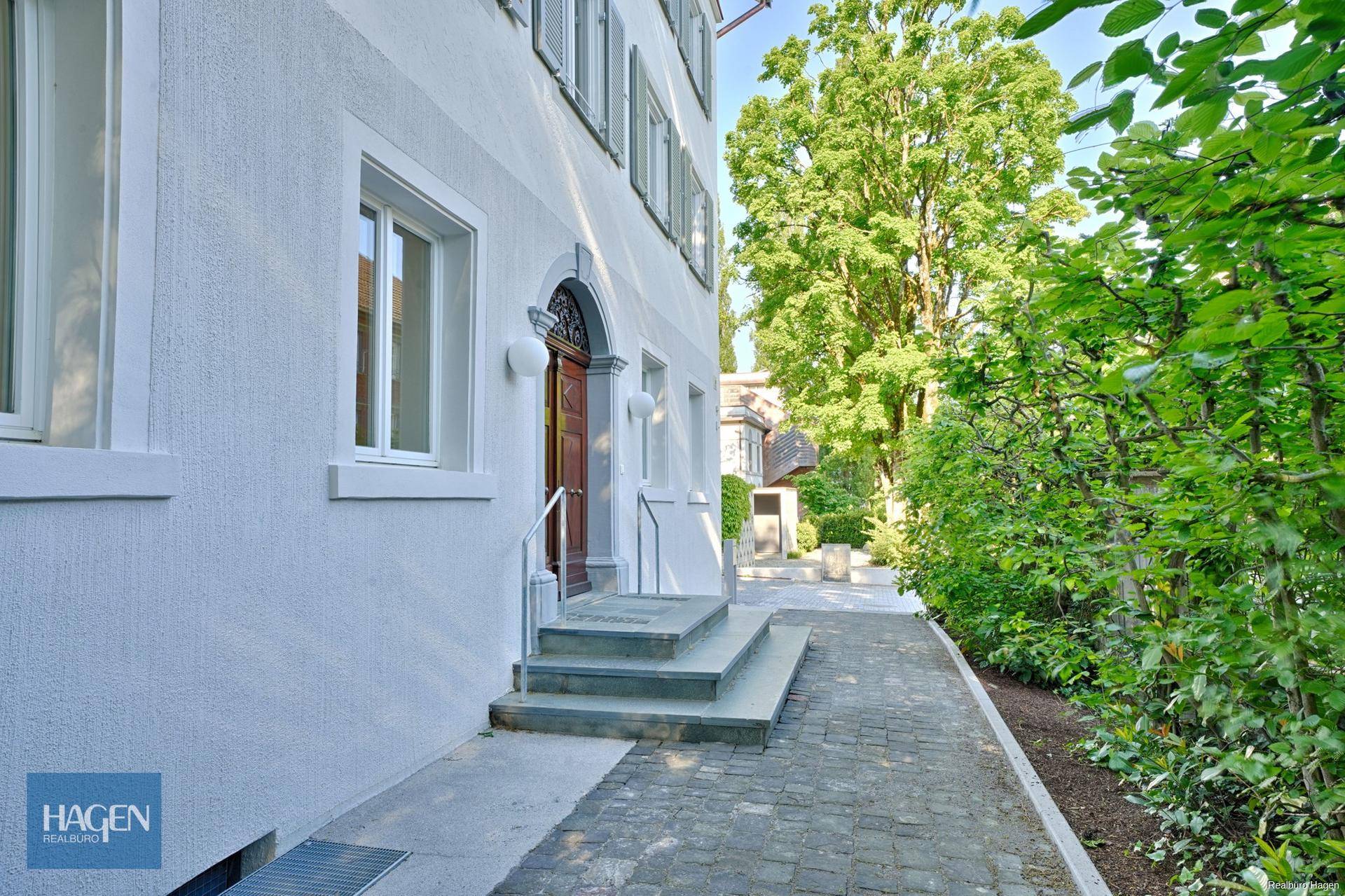 Eingangsbereich des Gebäudes mit einer Holztür, Steinstufen und einem gepflasterten Weg, umgeben von Hecken.