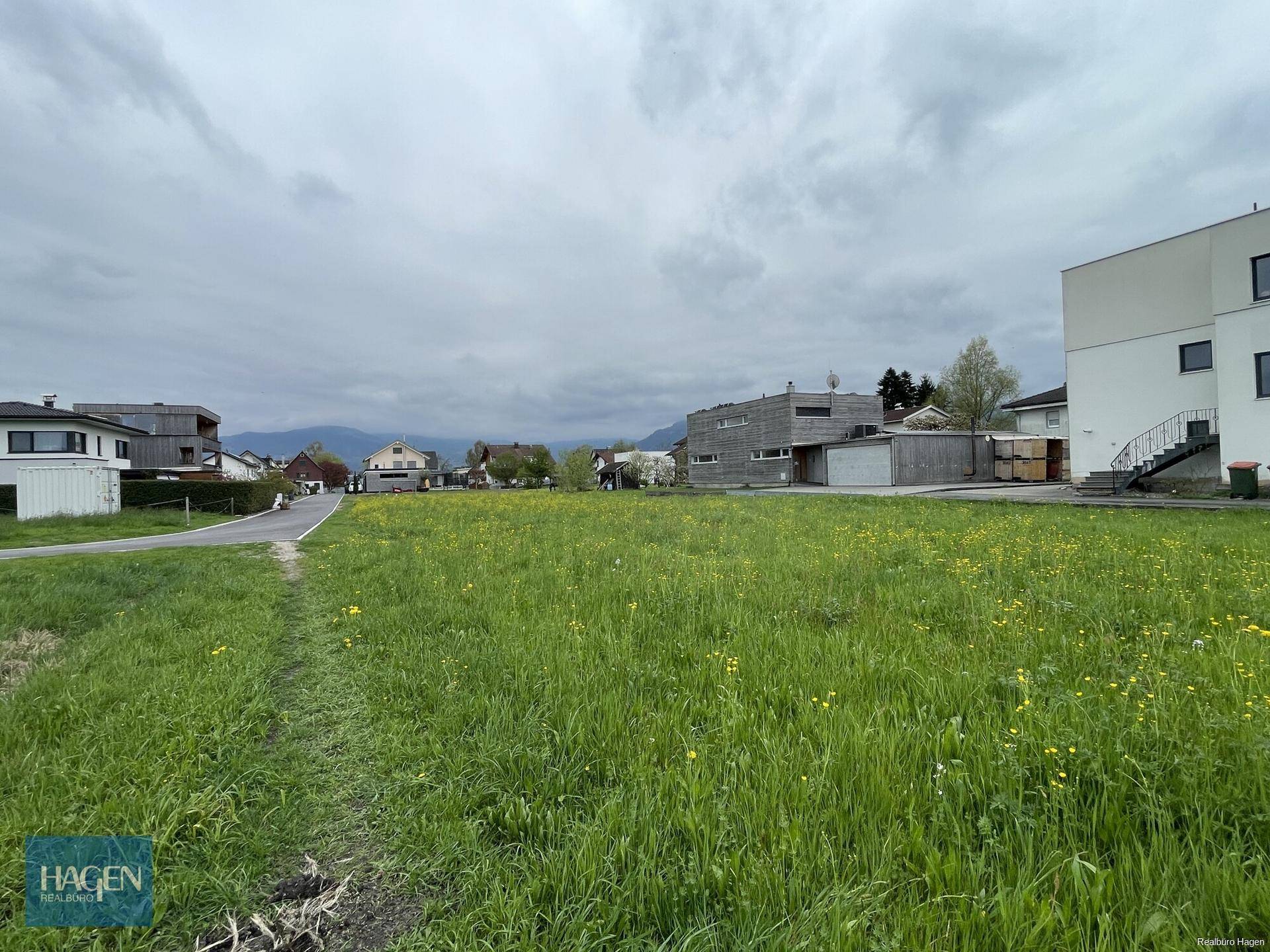 Unbebautes Grundstück mit Wiesen und einem Pfad, umgeben von Wohnbebauung und Bergen im Hintergrund.