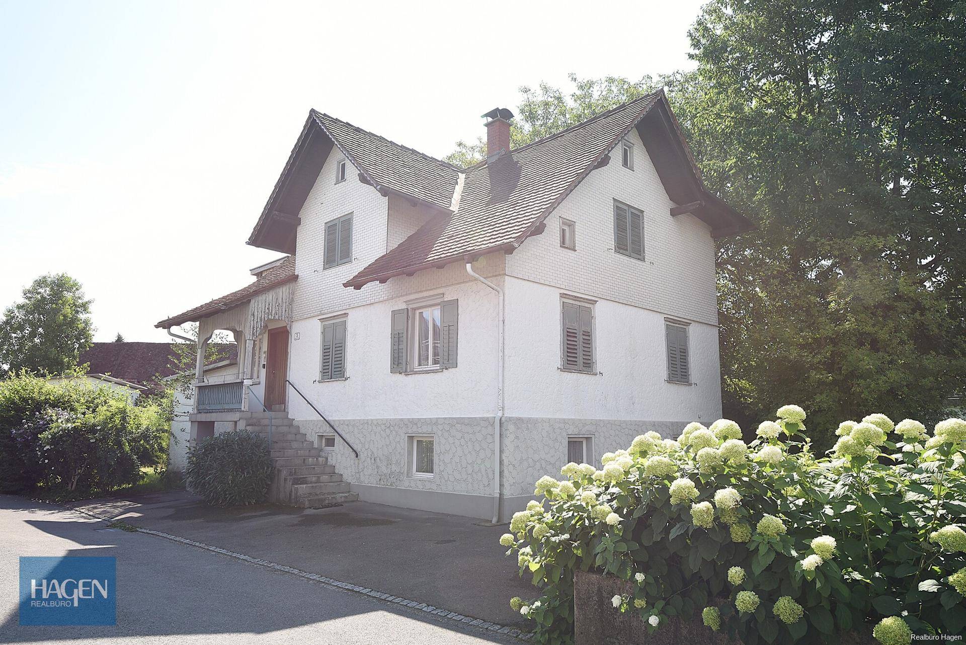 Frontansicht des Einfamilienhauses mit weißer Fassade, grünen Fensterläden und blühenden Sträuchern.