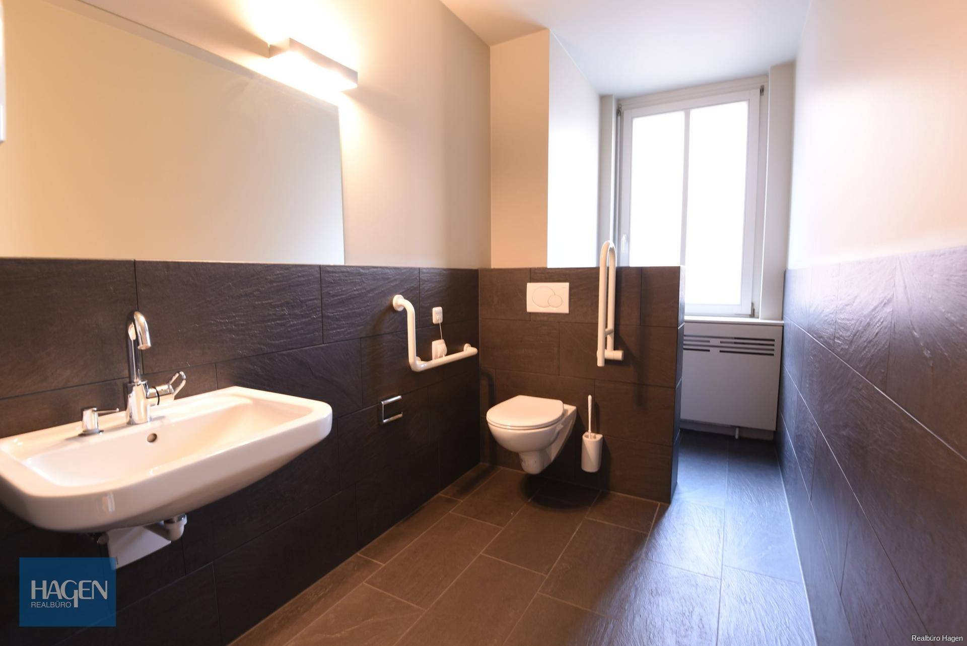 Barrierefreies Badezimmer mit WC, Waschbecken, Haltegriffen und Fenster für natürliches Tageslicht.