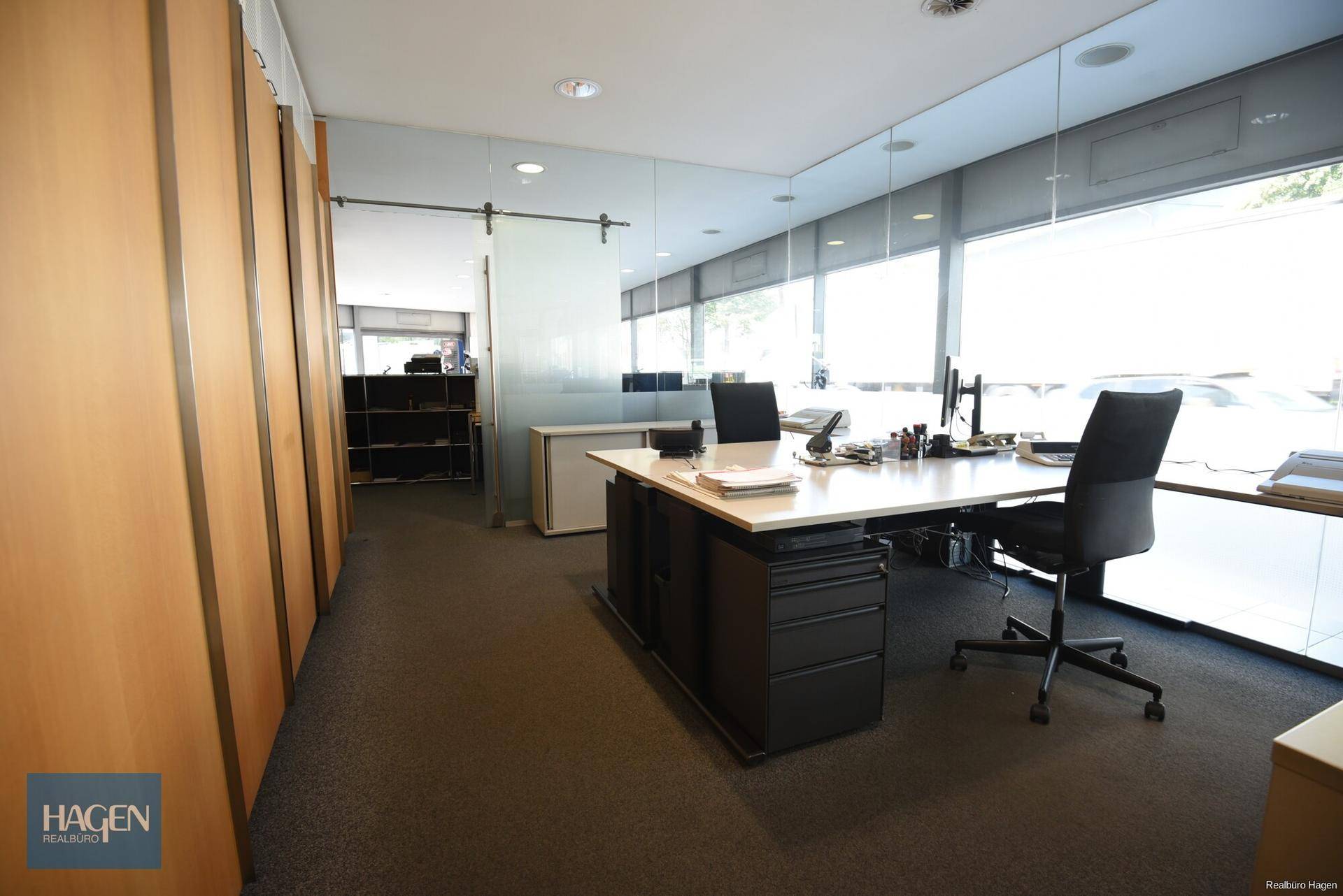 Modernes Büro mit großem Schreibtisch, Bürostühlen und raumhohen Fenstern, die viel Tageslicht hereinlassen.