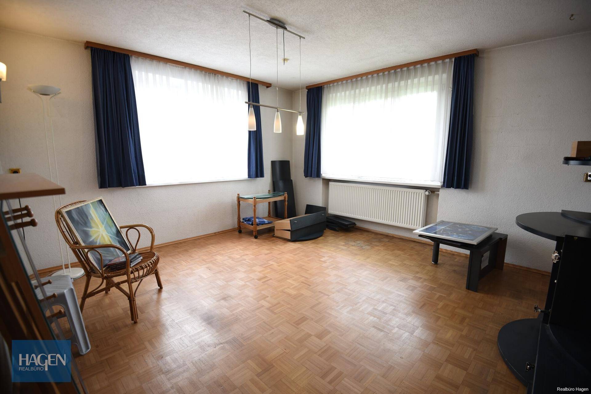 Geräumiges Wohnzimmer mit Parkettboden, großen Fenstern und blauen Vorhängen.