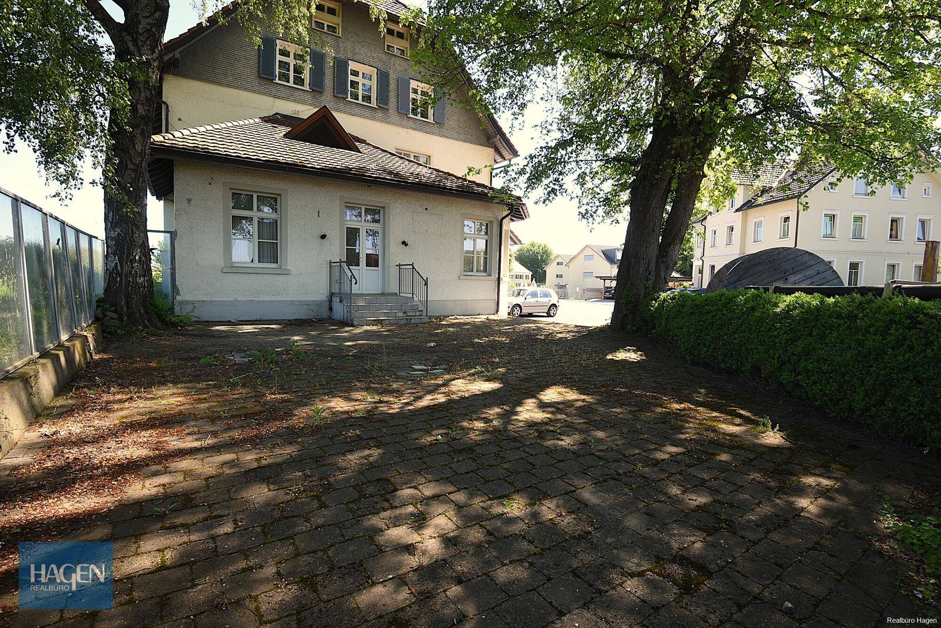 Gepflasterter Außenbereich mit Bäumen und Grünflächen hinter dem Gebäude, ideal für Entspannung.