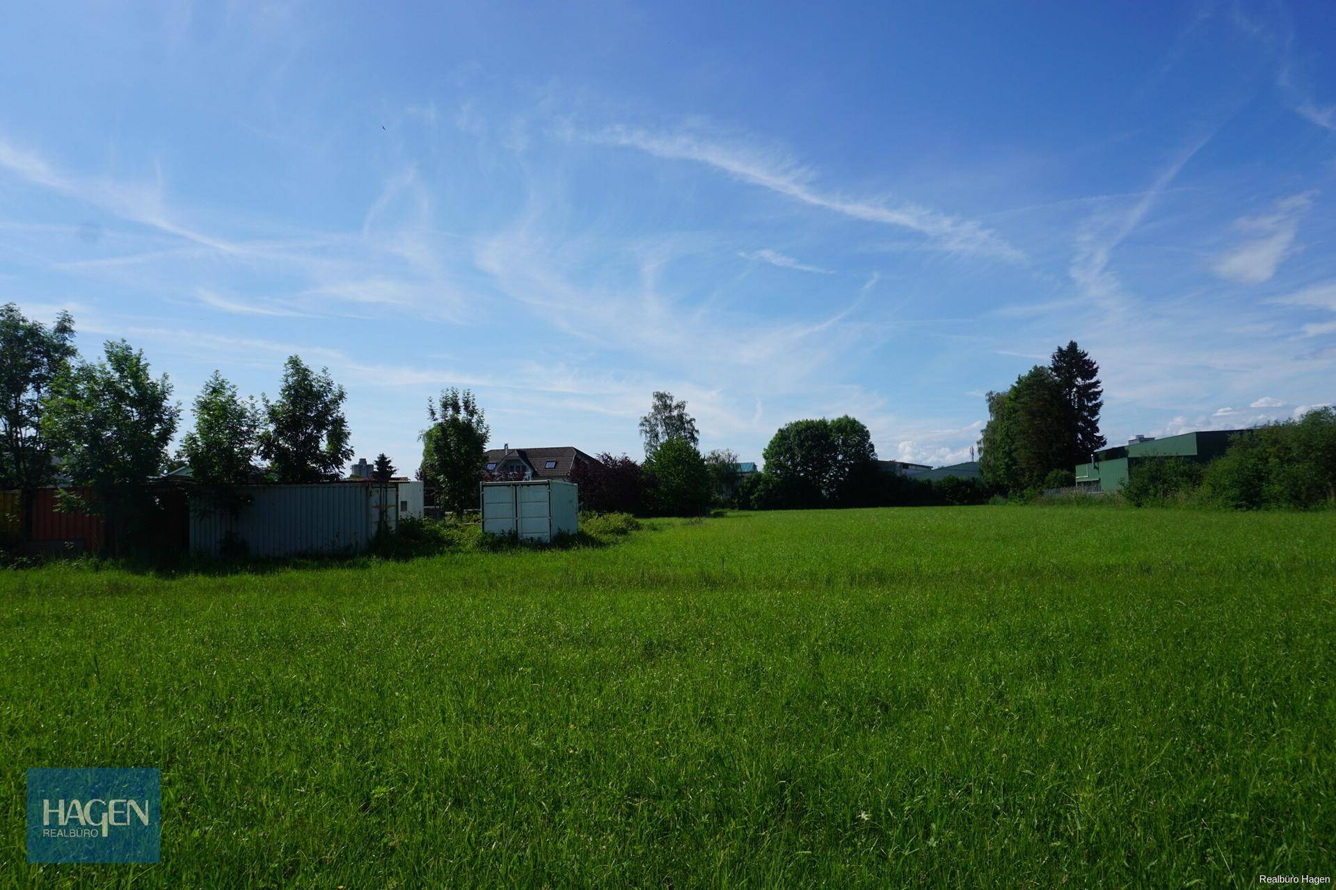 Grüne Wiese mit kleinen Schuppen und Bäumen unter strahlend blauem Himmel, die eine offene Fläche darstellt.