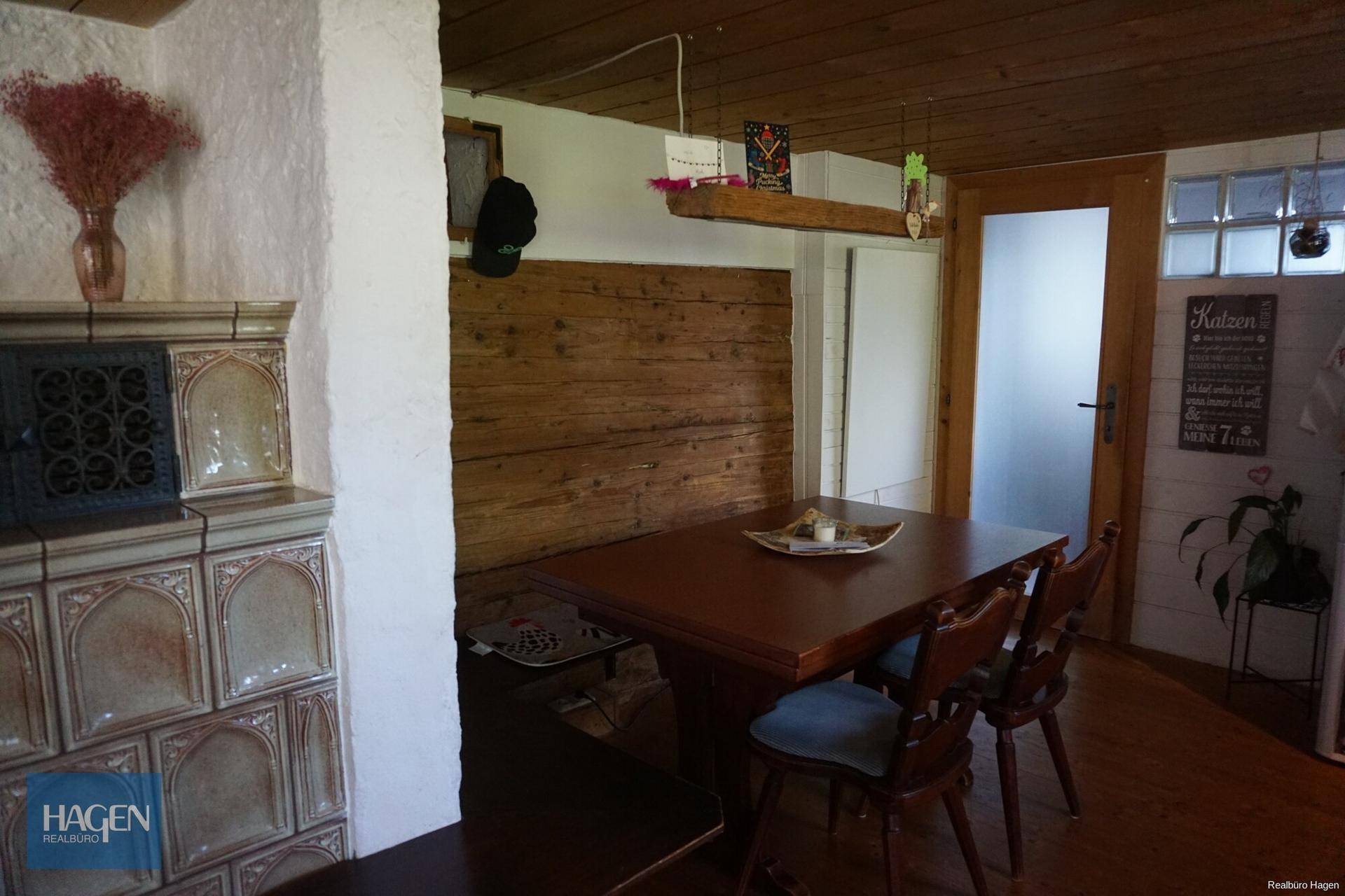 Rustikaler Essbereich mit Holztisch, Sitzbank, Kachelofen und Holzvertäfelung an Wänden und Decke.