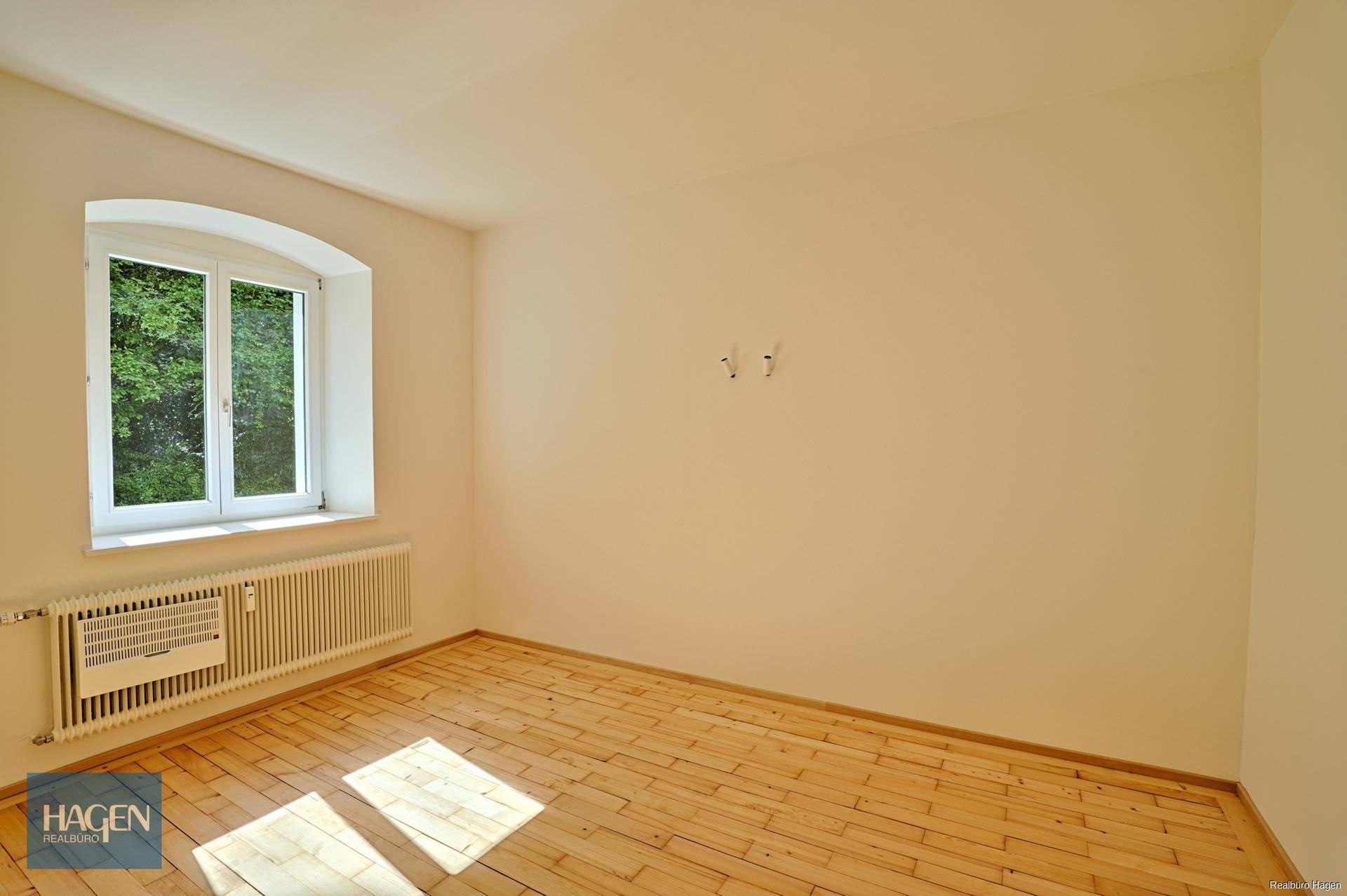 Leeres Zimmer mit Dielenboden, weißen Wänden und einem Fenster mit Blick ins Grüne.
