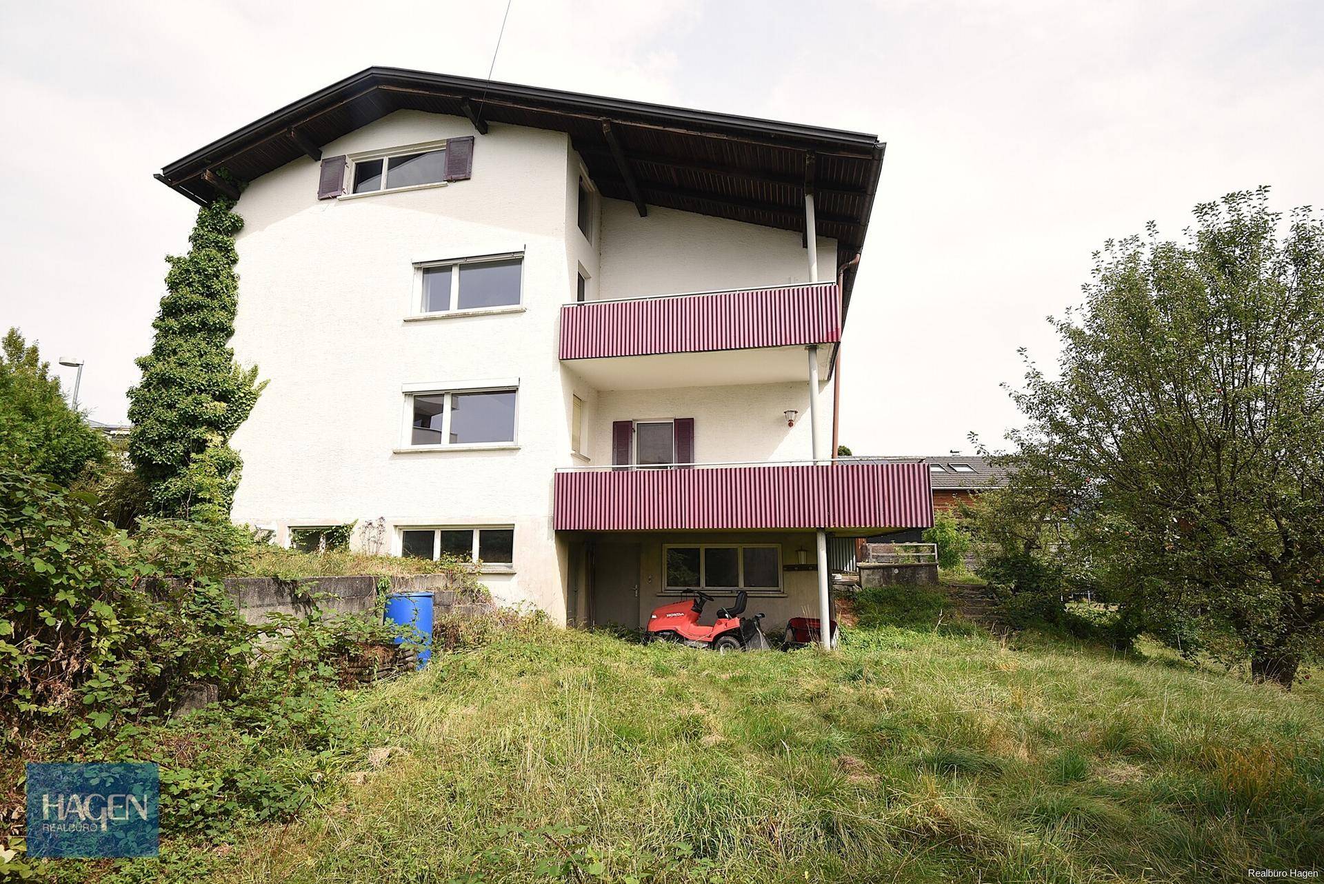 Seitenansicht des Hauses mit zwei Balkonen und einem ungepflegten Gartenbereich im Vordergrund.