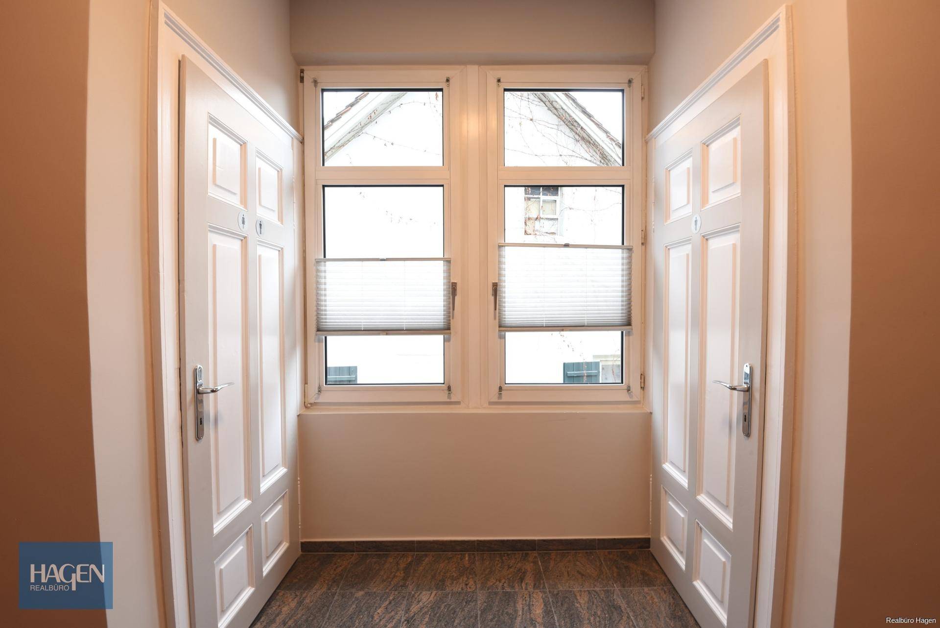 Heller Flur mit zwei weißen Türen, einem Fenster und dunklem Fliesenboden.