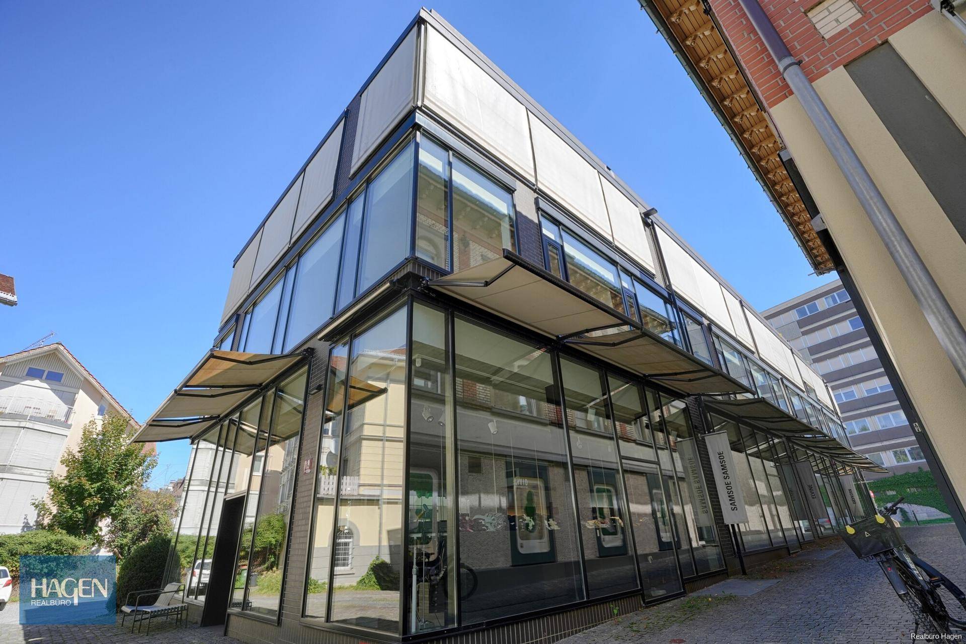 Moderne Geschäftsfassade mit großen Glasflächen und Sonnenschutz unter blauem Himmel.