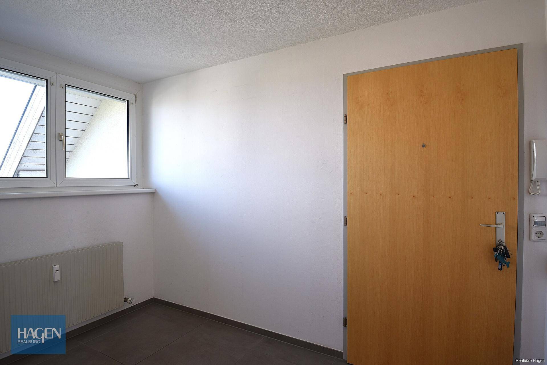 Eingangsbereich mit Holztür, Fliesenboden, Heizkörper und Fenster für Tageslicht.