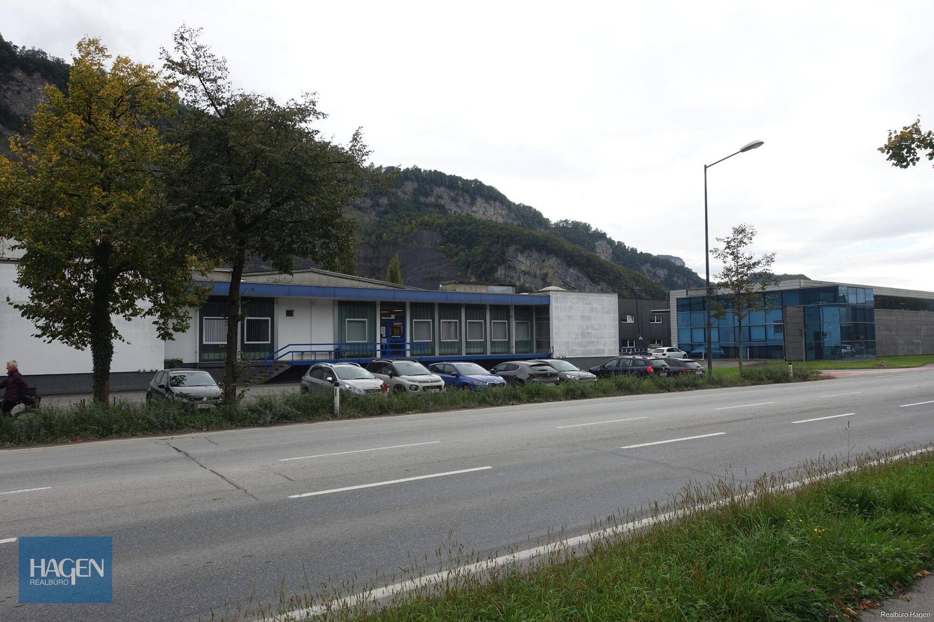 Gebäudeansicht von der Straße aus mit geparkten Autos und Bergen im Hintergrund unter bewölktem Himmel.