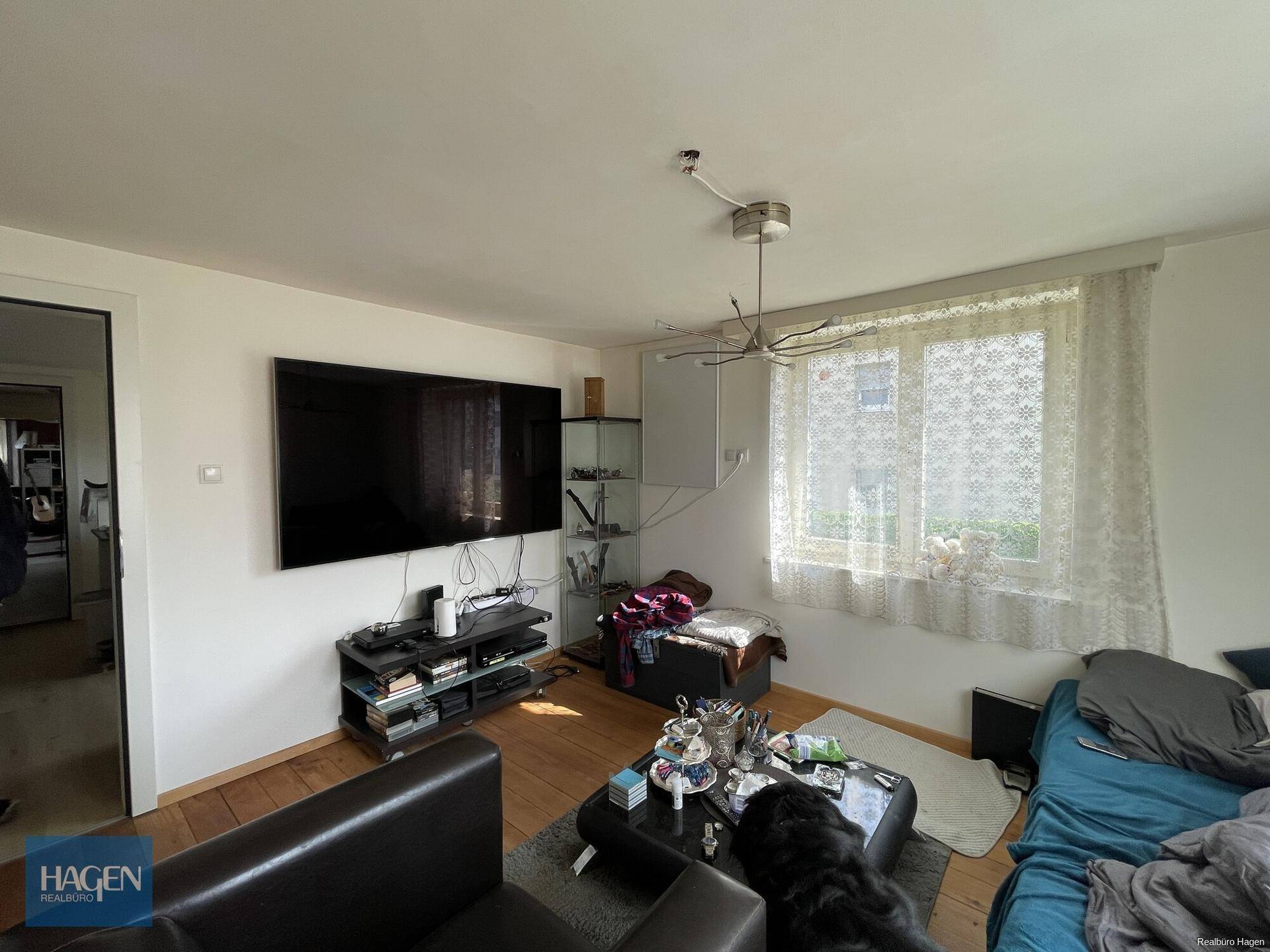 Gemütliches Wohnzimmer mit großem Wandfernseher, Holzboden und viel Tageslicht.