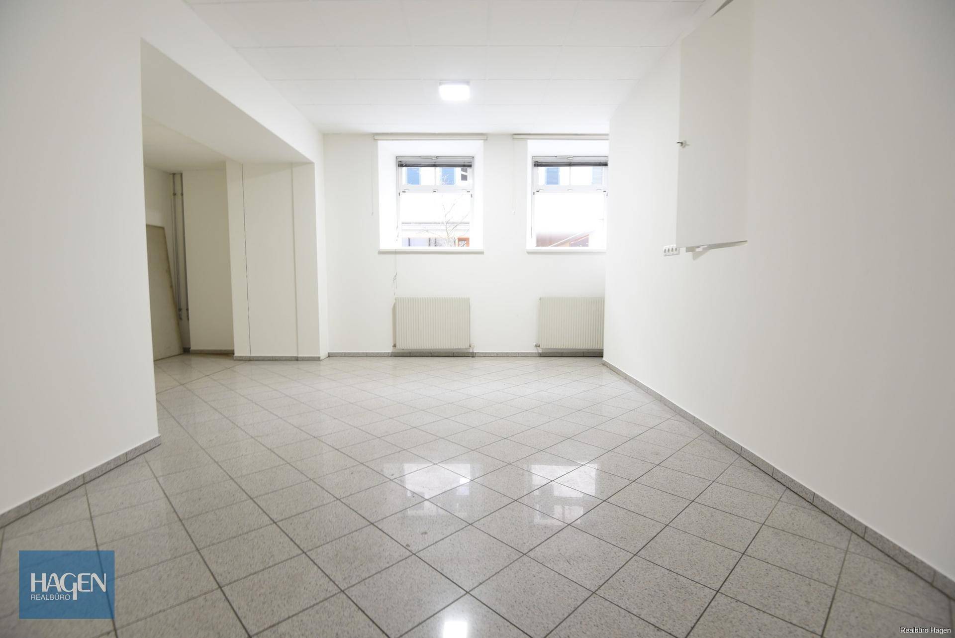Leerer Raum mit zwei Fenstern, Heizkörpern und Fliesenboden, ideal für Büro oder Lager.
