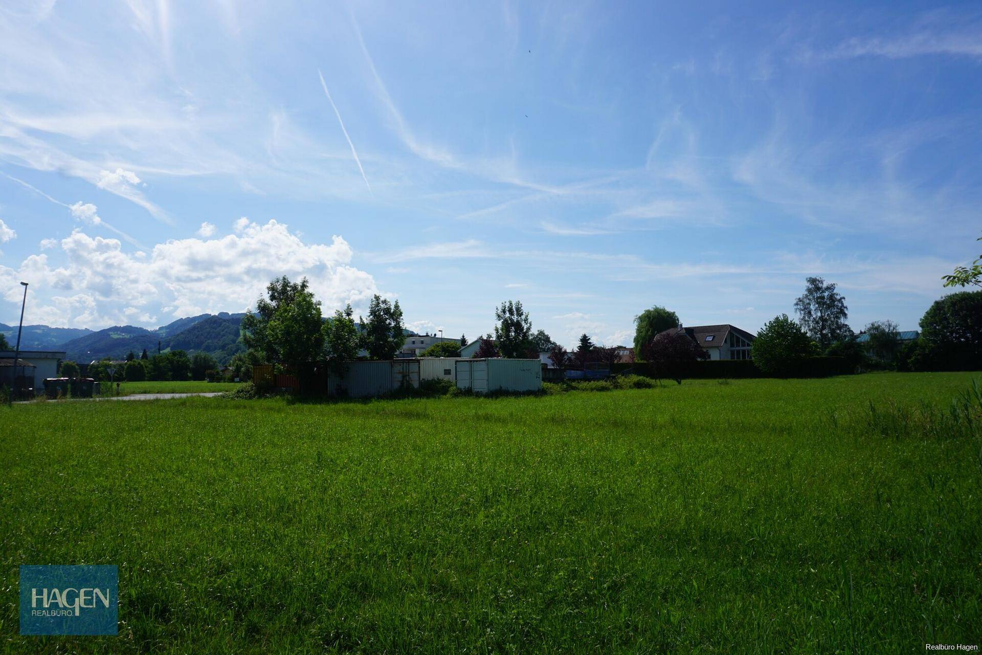 Weite grüne Fläche mit Schuppen und einem Panoramablick auf die Berge im Hintergrund unter blauem Himmel.