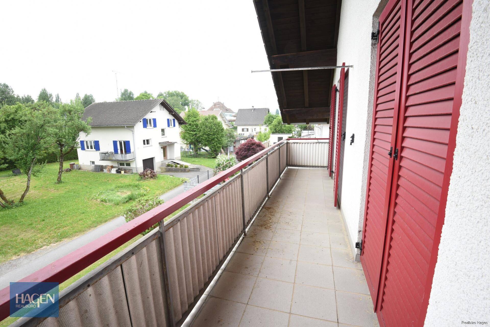 Langer Balkon mit Fliesenboden und roten Fensterläden, bietet Blick auf die Umgebung.