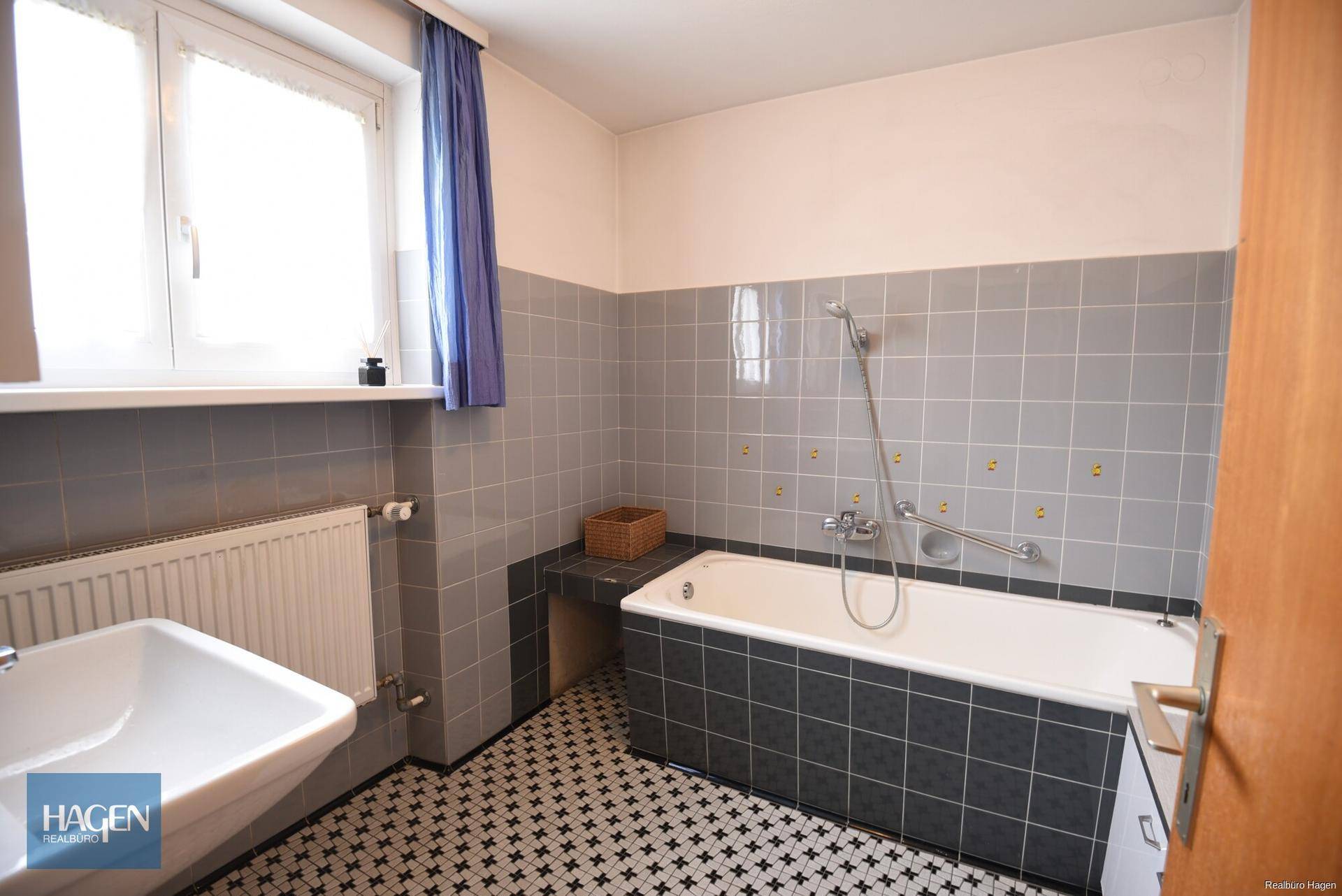 Badezimmer mit Badewanne, Duscharmatur, Haltegriffen und grauen Fliesen.