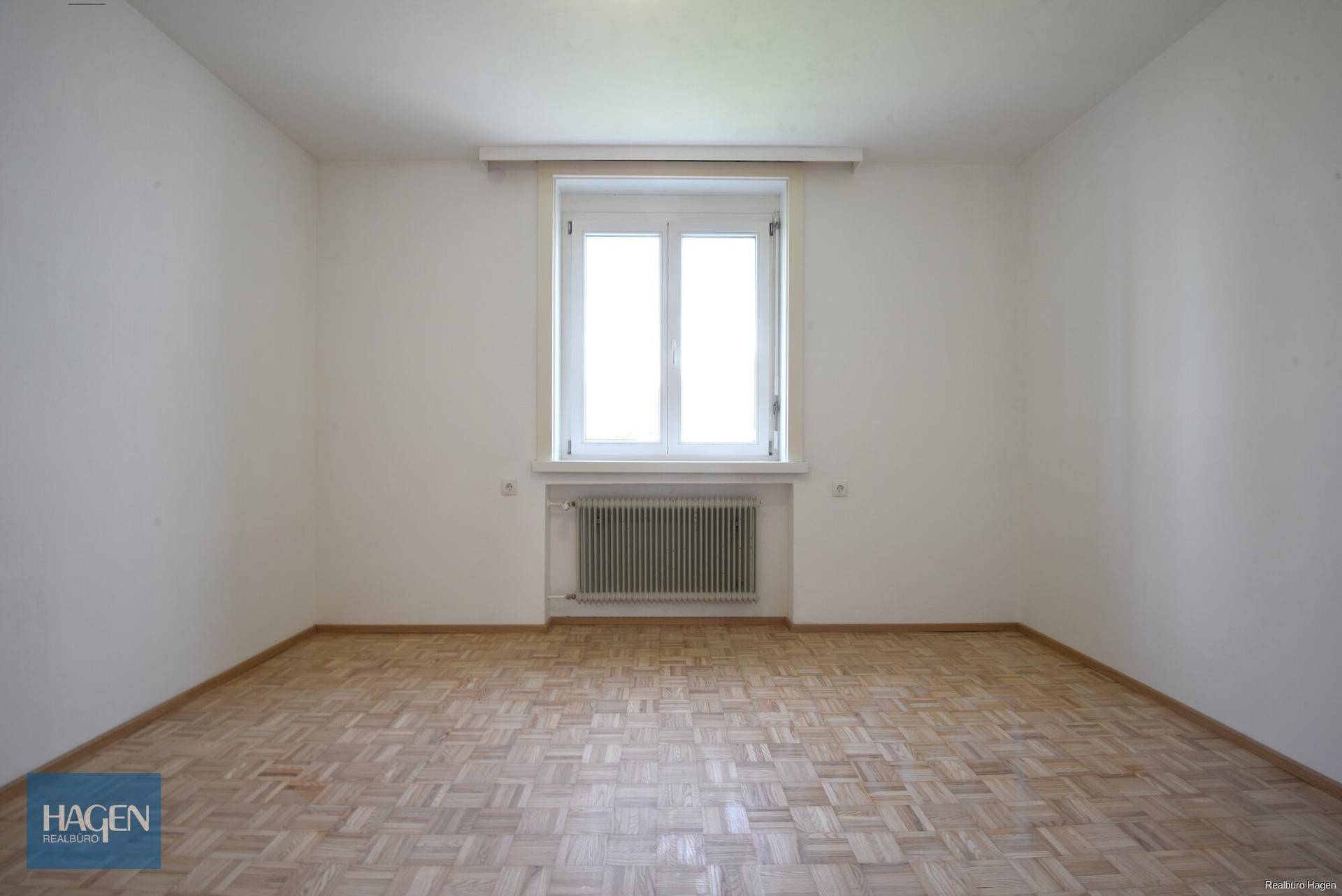 Helles Zimmer mit Parkettboden und einem großen Fenster, das viel Tageslicht hereinlässt.