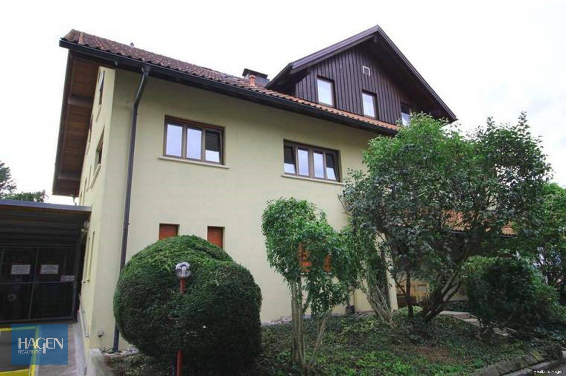 Ansicht eines älteren Gebäudes mit beiger Fassade und braunem Dach, umgeben von Bäumen und Sträuchern, mit einer Zufahrt auf der linken Seite.