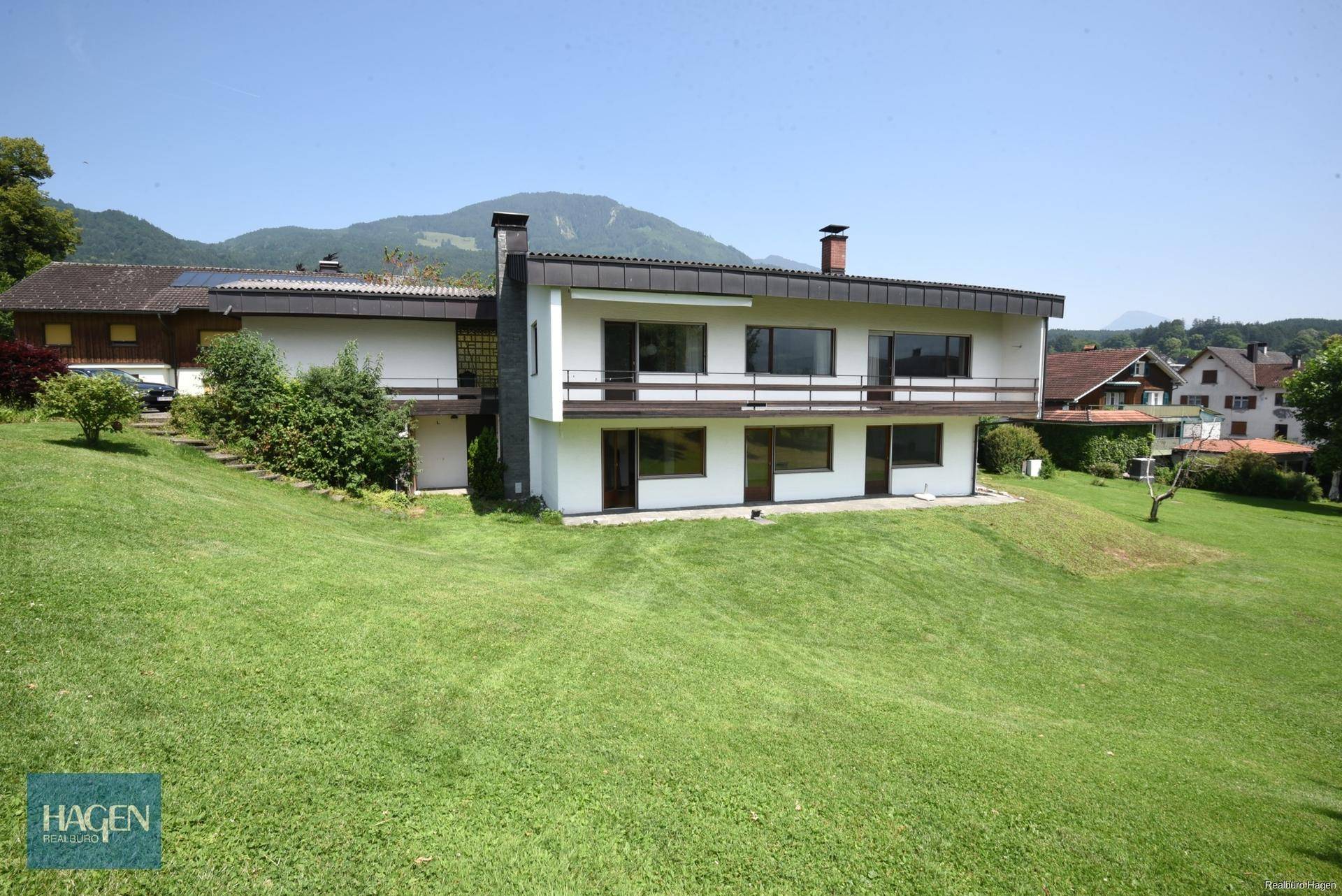 Weitläufiges Grundstück mit Einfamilienhaus und Bergblick unter blauem Himmel.