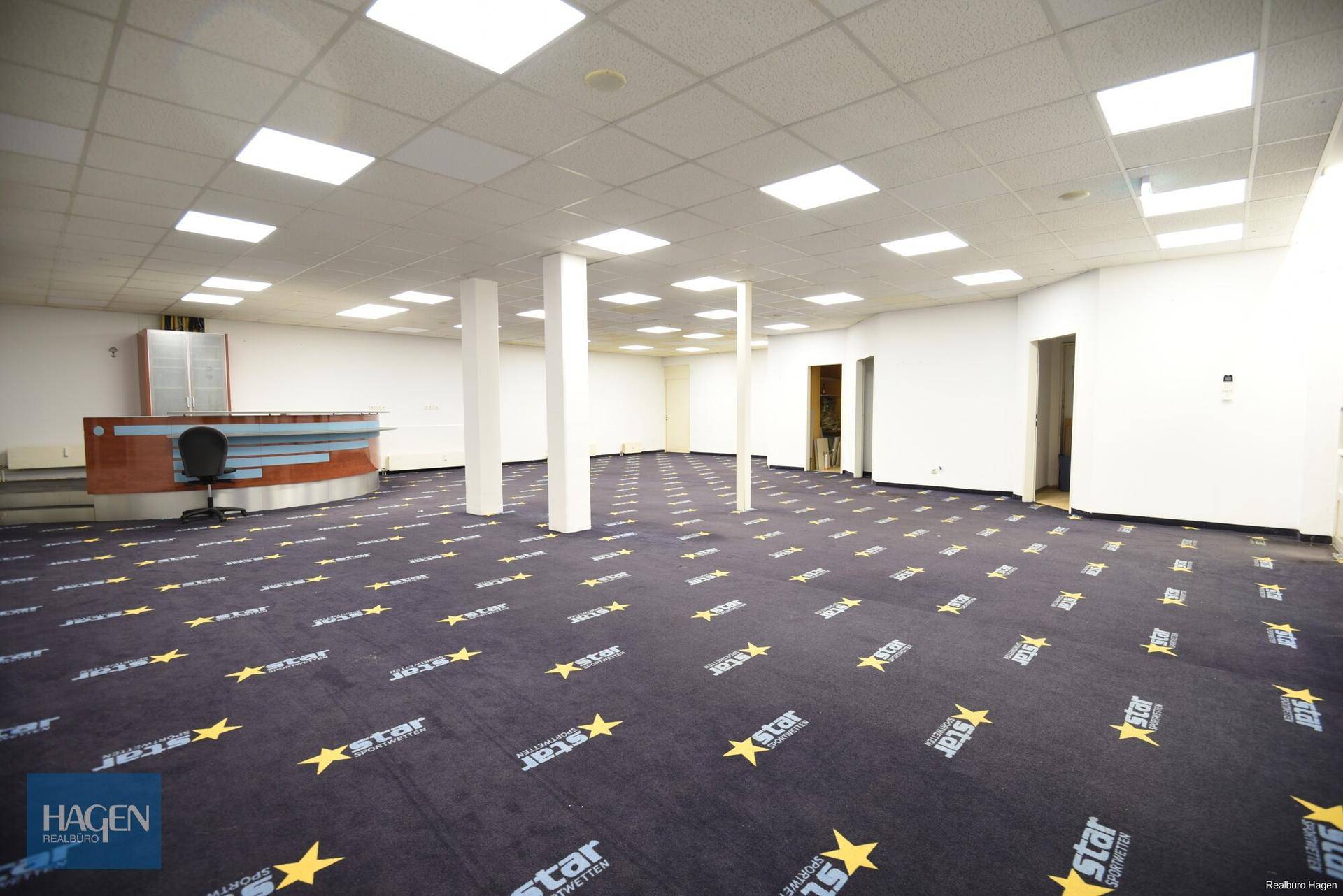 Großer, leerer Gewerberaum mit auffälligem Teppichboden, Empfangstheke und Säulen, beleuchtet von Deckenleuchten.