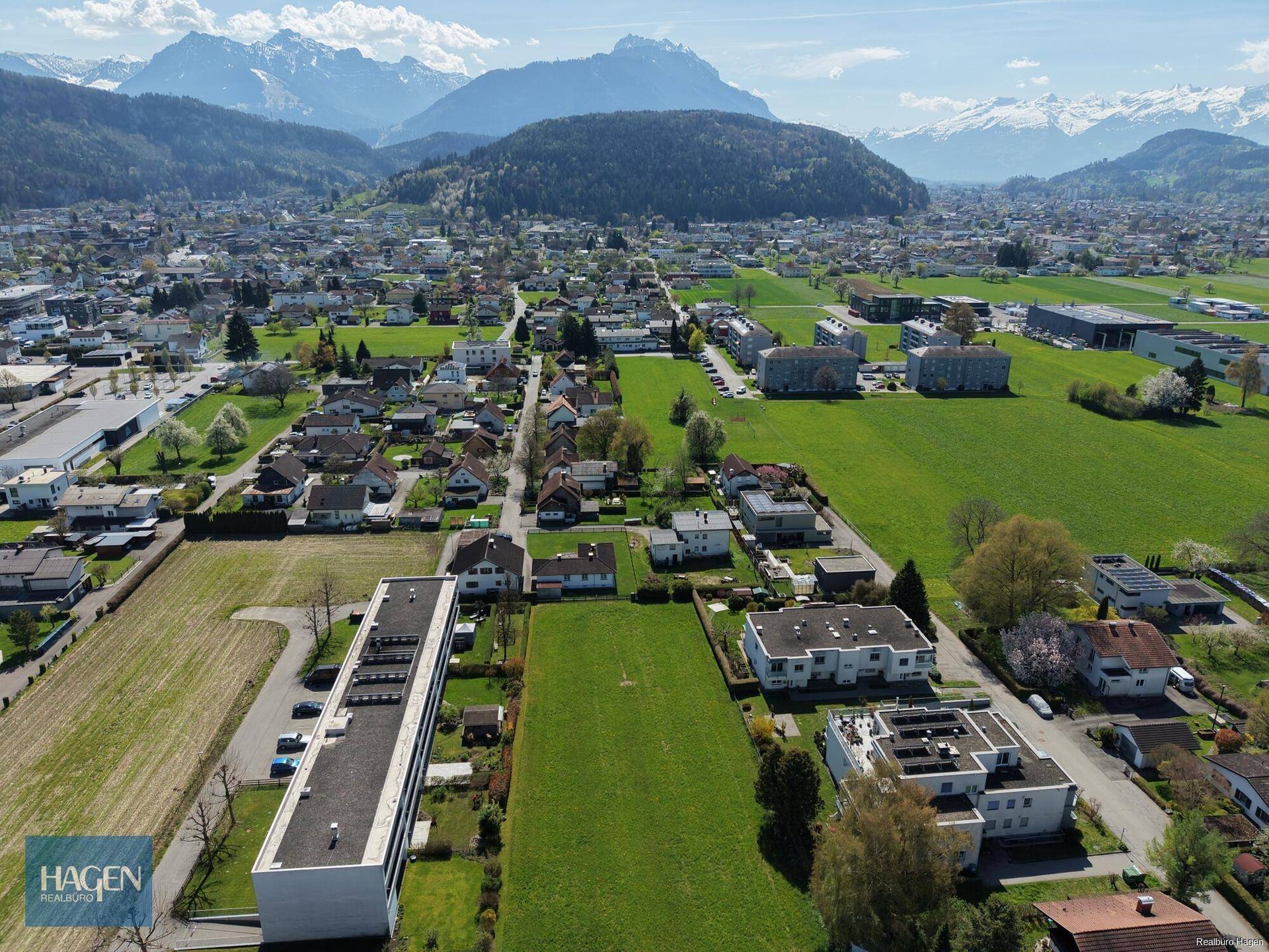 Weitläufige Luftaufnahme der Wohngegend mit grünen Feldern und majestätischen Bergen im Hintergrund.