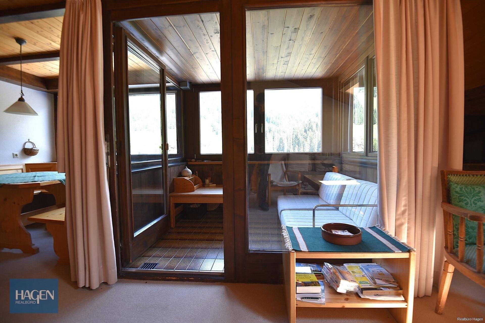 Blick vom Wohnzimmer in einen hellen Wintergarten mit Holzboden und großen Fenstern, umgeben von Natur.