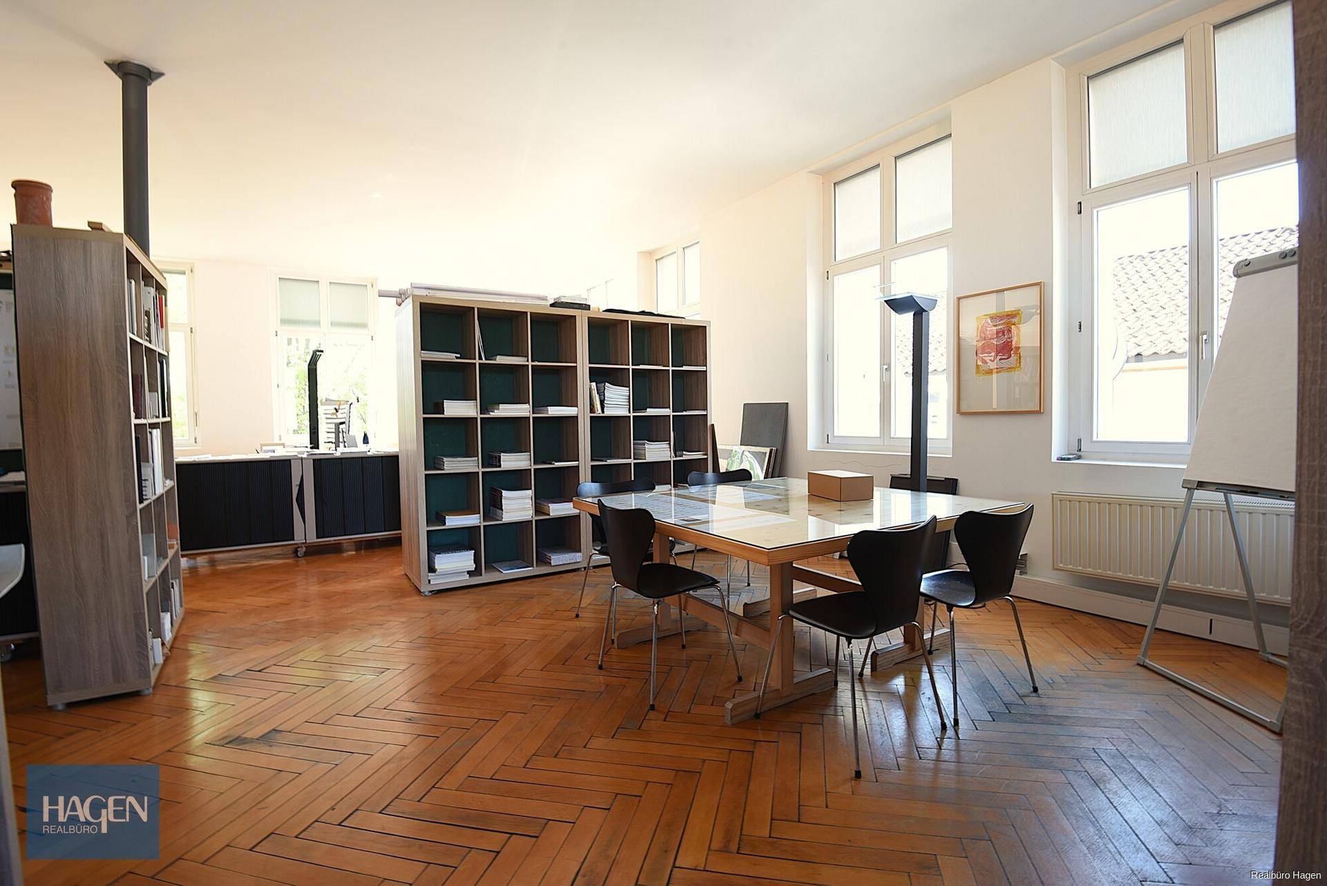 Heller Besprechungsraum mit Fischgrätparkett, großen Fenstern und Bücherregalen, ideal für Teamarbeit.