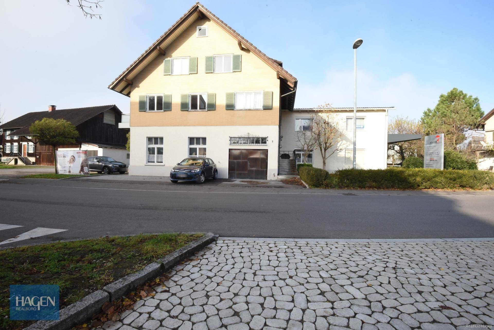 Vorderansicht des Gebäudes mit Parkplätzen und angrenzender Straße, umgeben von Bäumen und Büschen.