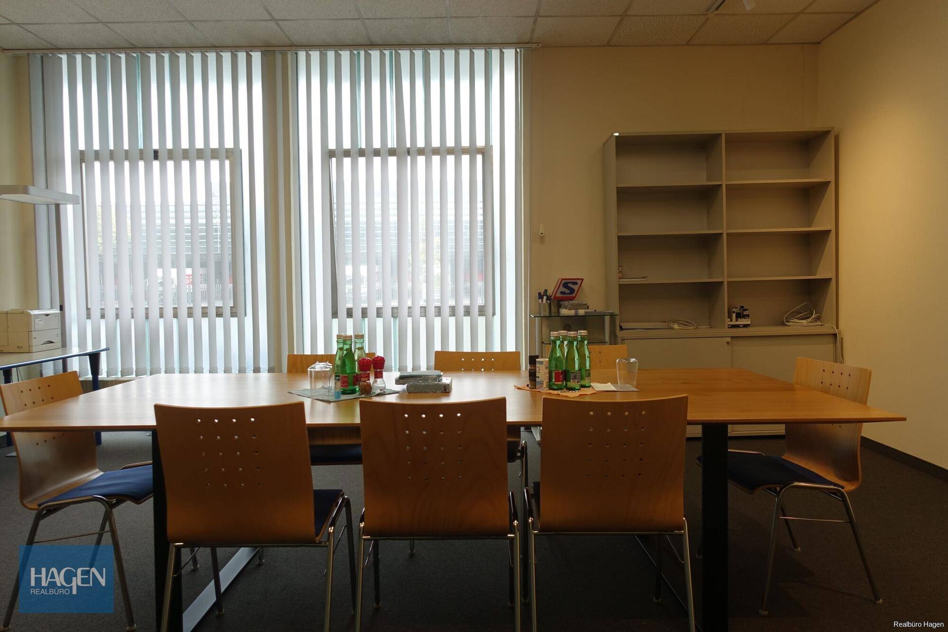 Besprechungsraum mit großem Holztisch, mehreren Stühlen und vertikalen Jalousien an den Fenstern.