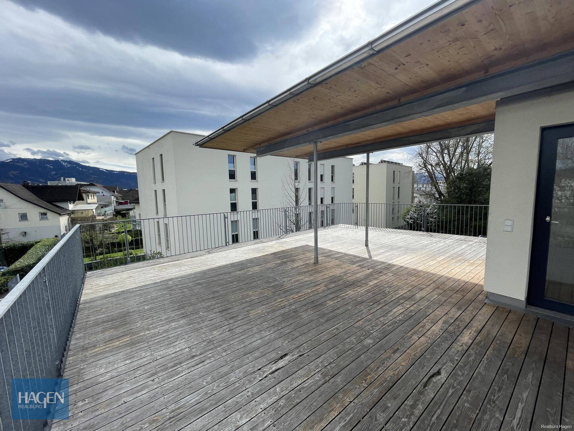 Große Holzterrasse mit Metallgeländer und Blick auf die umliegenden Gebäude und Berge.