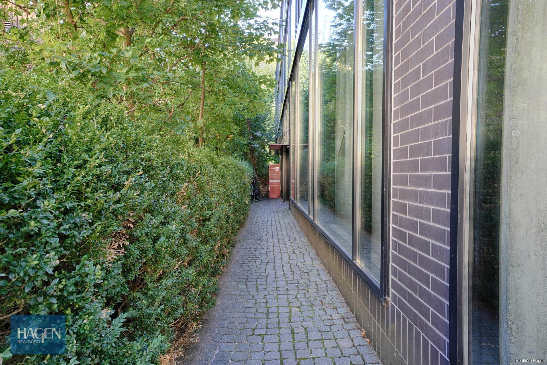 Gepflasterter Weg entlang der Gebäudeseite mit Hecken und Bäumen.