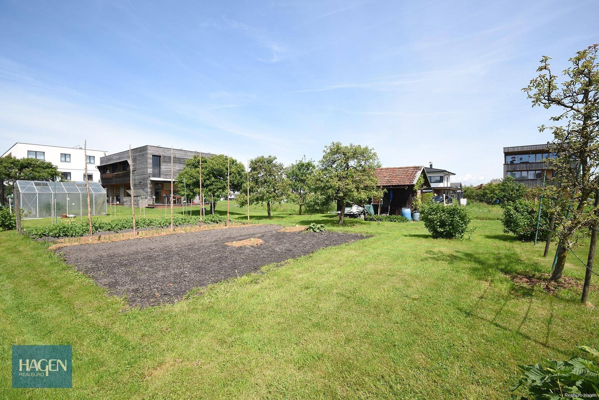 Garten mit einem vorbereiteten Beet, einem Gewächshaus und verschiedenen Bäumen, ideal für den Anbau von Gemüse.