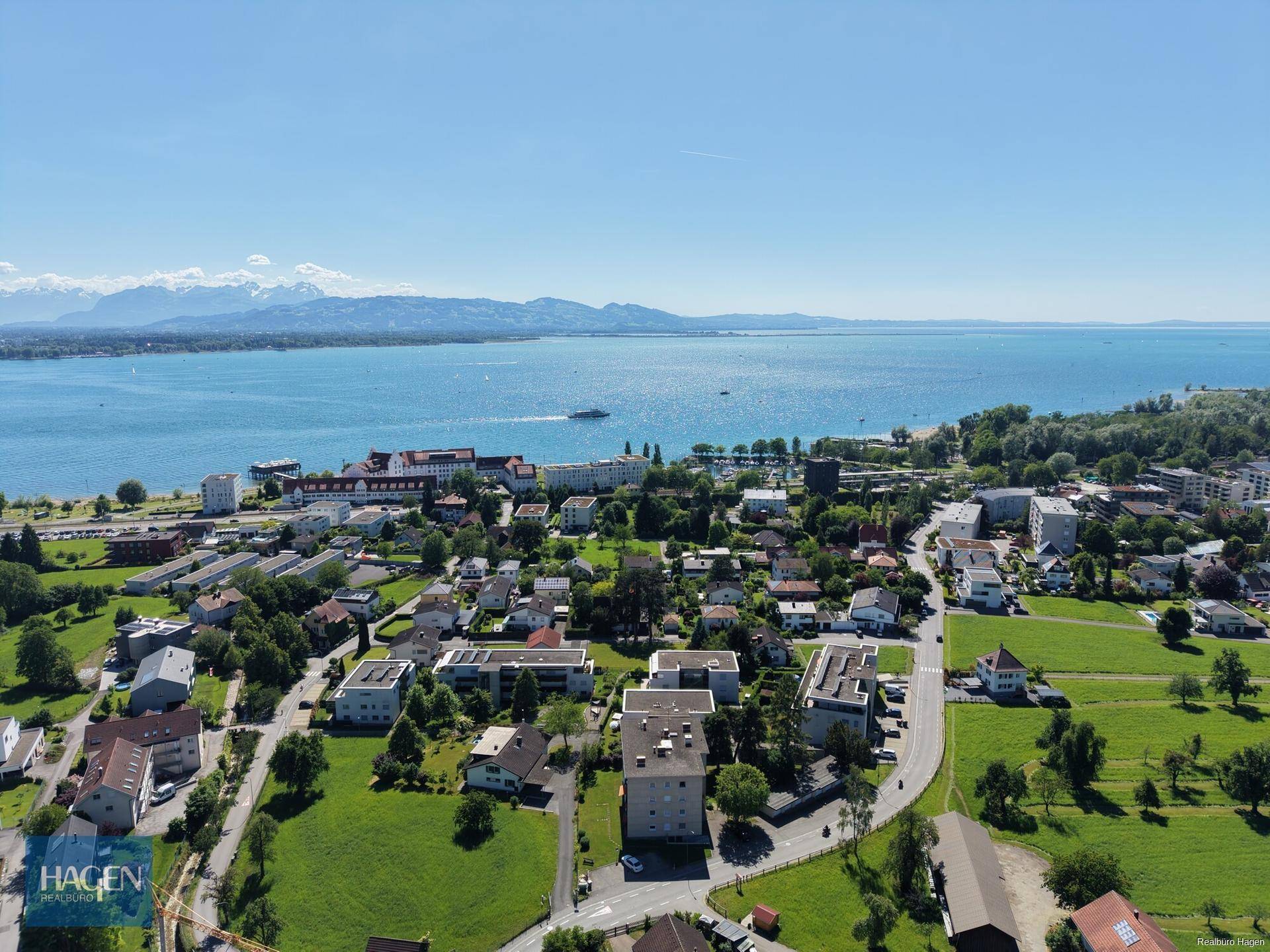 Panoramablick aus der Vogelperspektive auf eine Stadt am See mit Bergen im Hintergrund und grünen Wiesen.