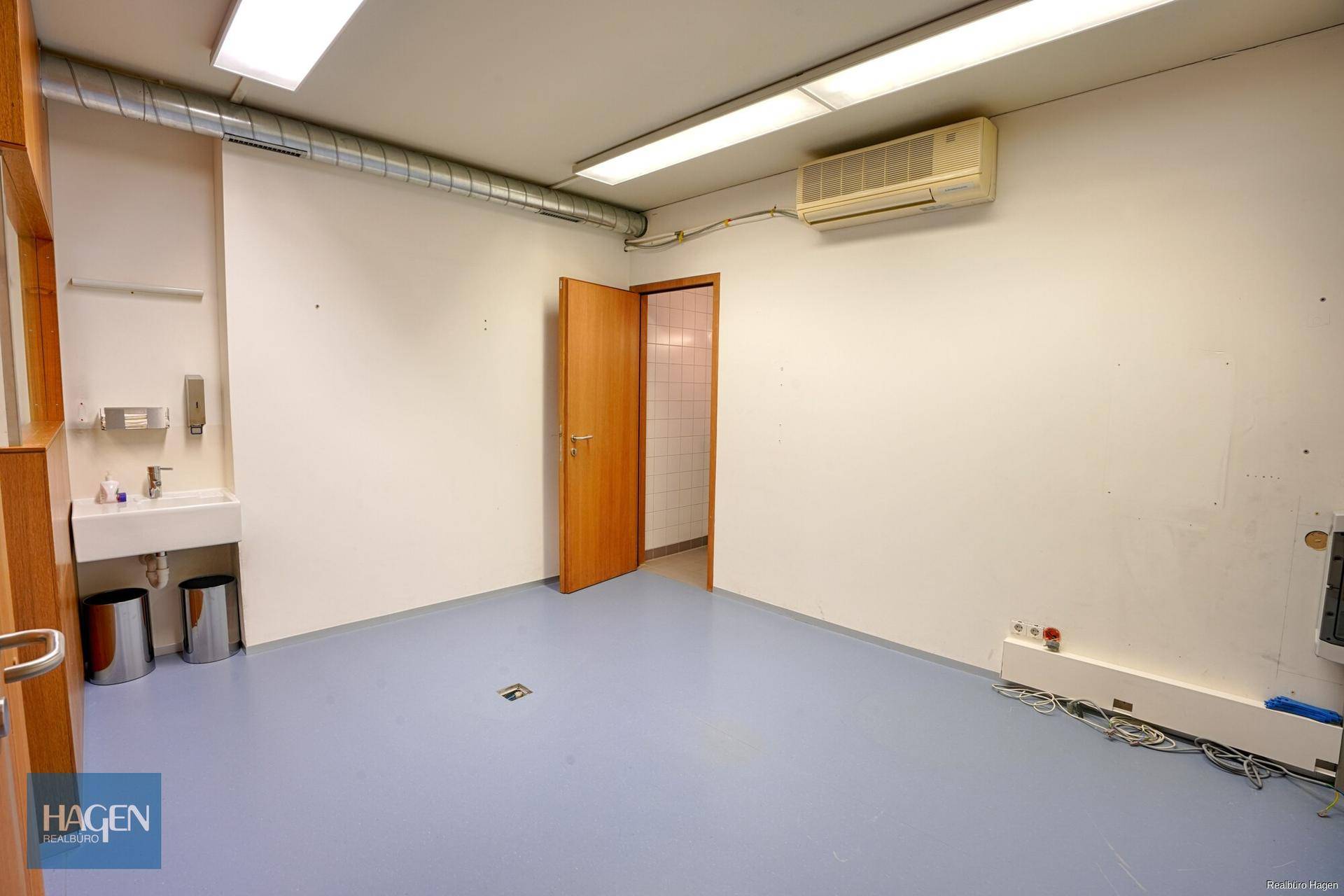 Leerer Raum mit blauer Bodenfläche, Klimaanlage und Zugang zu einem weiteren Bereich.