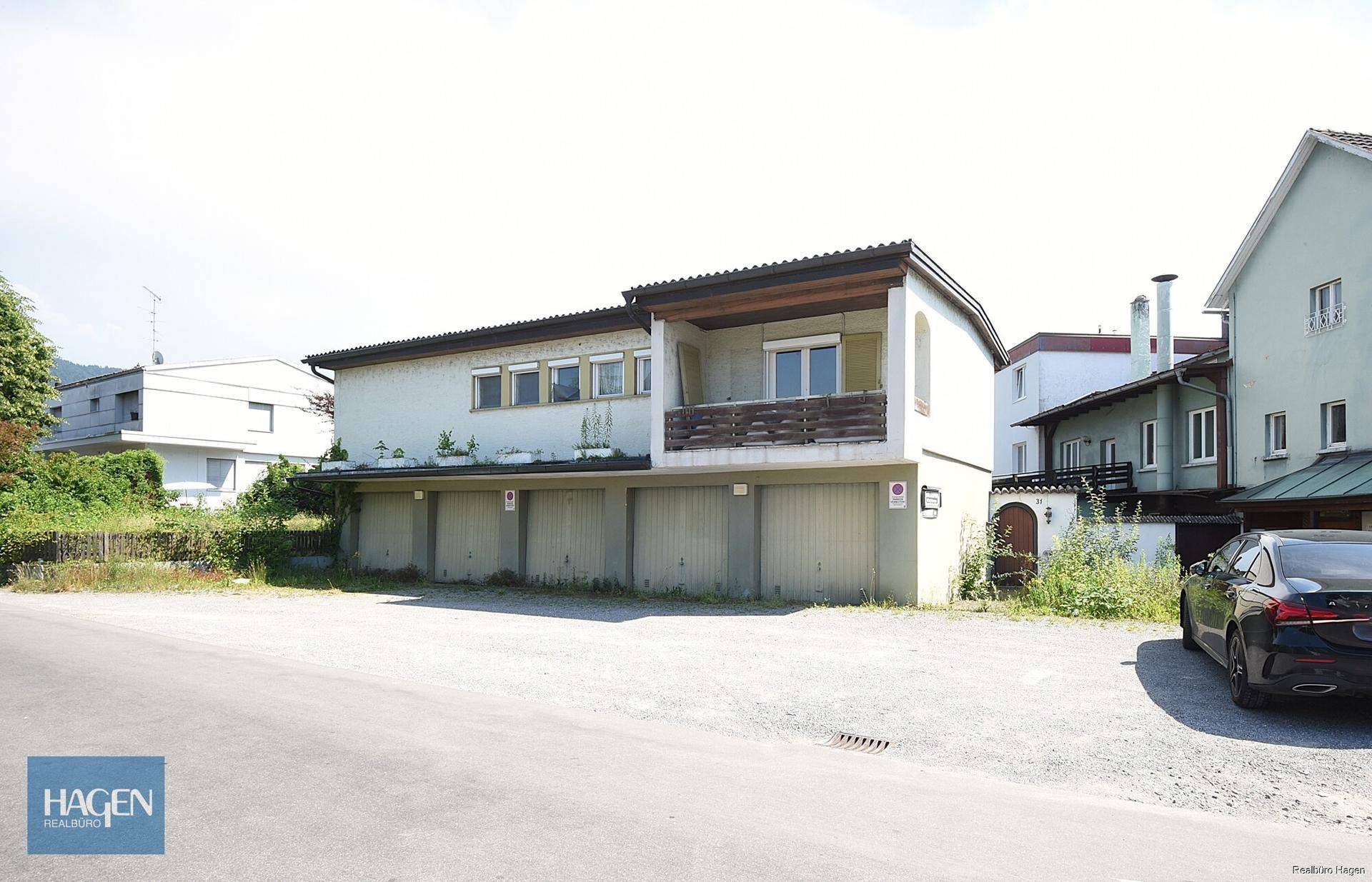 Gebäude mit mehreren Garagentoren im Erdgeschoss und einem Balkon im Obergeschoss, daneben ein Parkplatz.